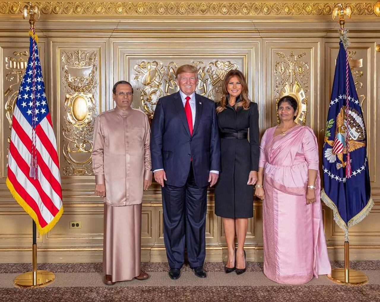 Donald Trump, flanked by Melania Trump, and two other people, stands in front of a gold wall with the American and Presidential flags