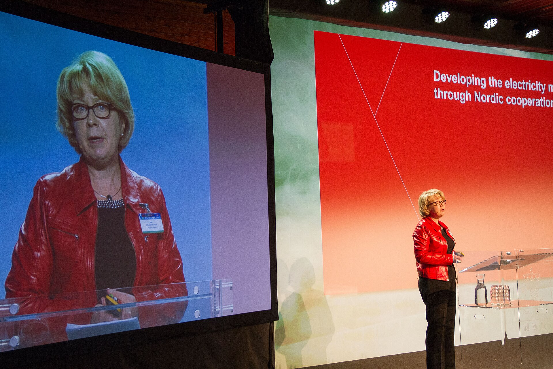 A woman in a red leather jacket and black pants stands on a stage in front of a large screen displaying her image. She is also visible on the right side of the image, standing in front of a glass podium with a large screen behind her showing a presentation slide about electricity and Nordic co.