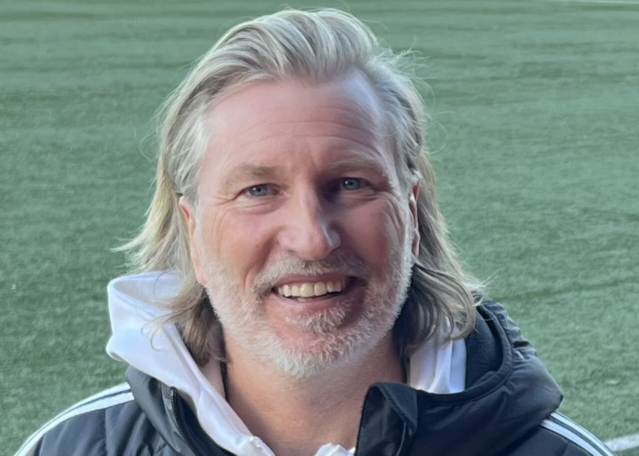 A smiling man with long blond hair and a short beard, wearing a black and white jacket, standing in front of a green field.