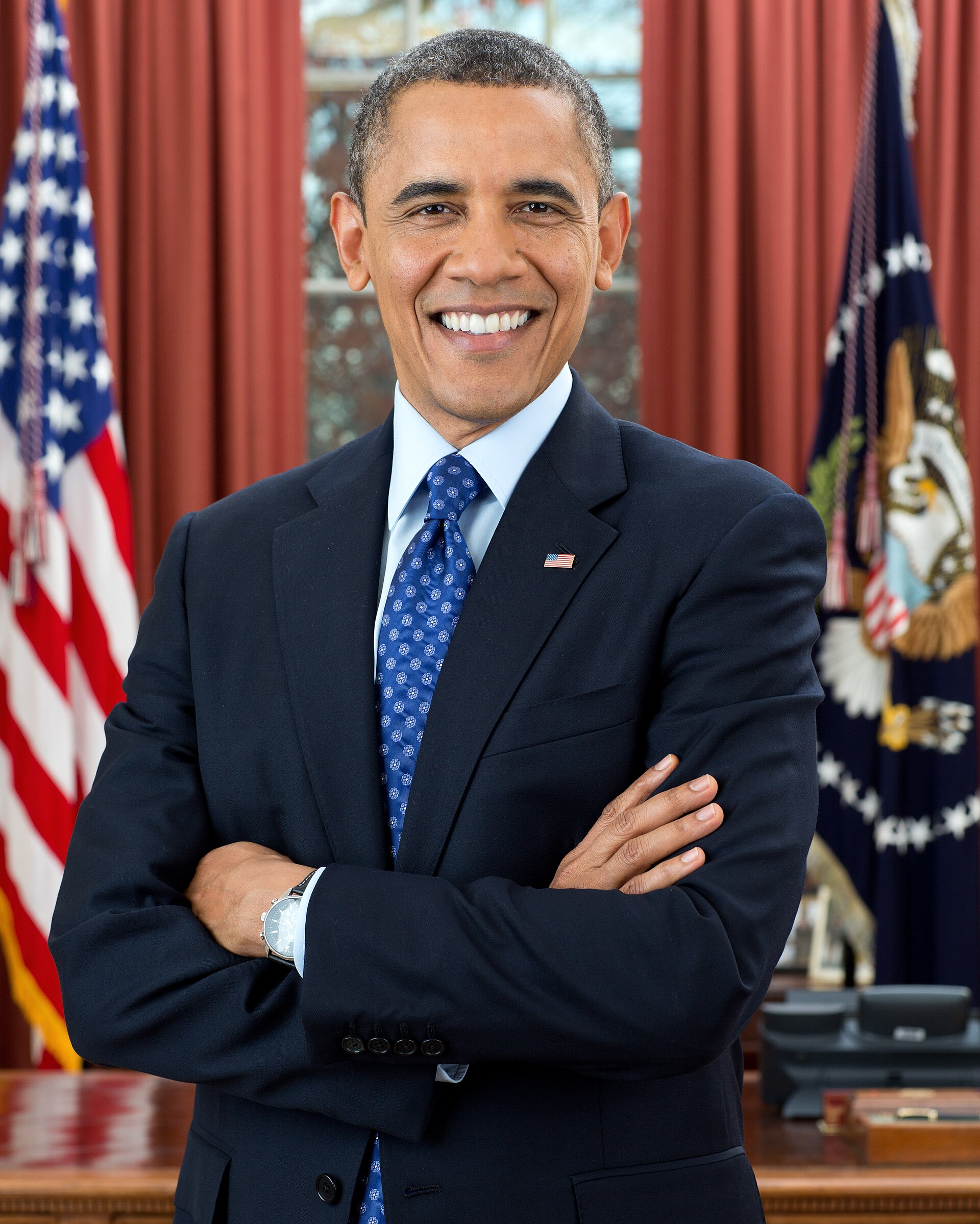 Barack Obama smiles with his arms crossed in the Oval Office, with the American flag and presidential seal flag behind him.
