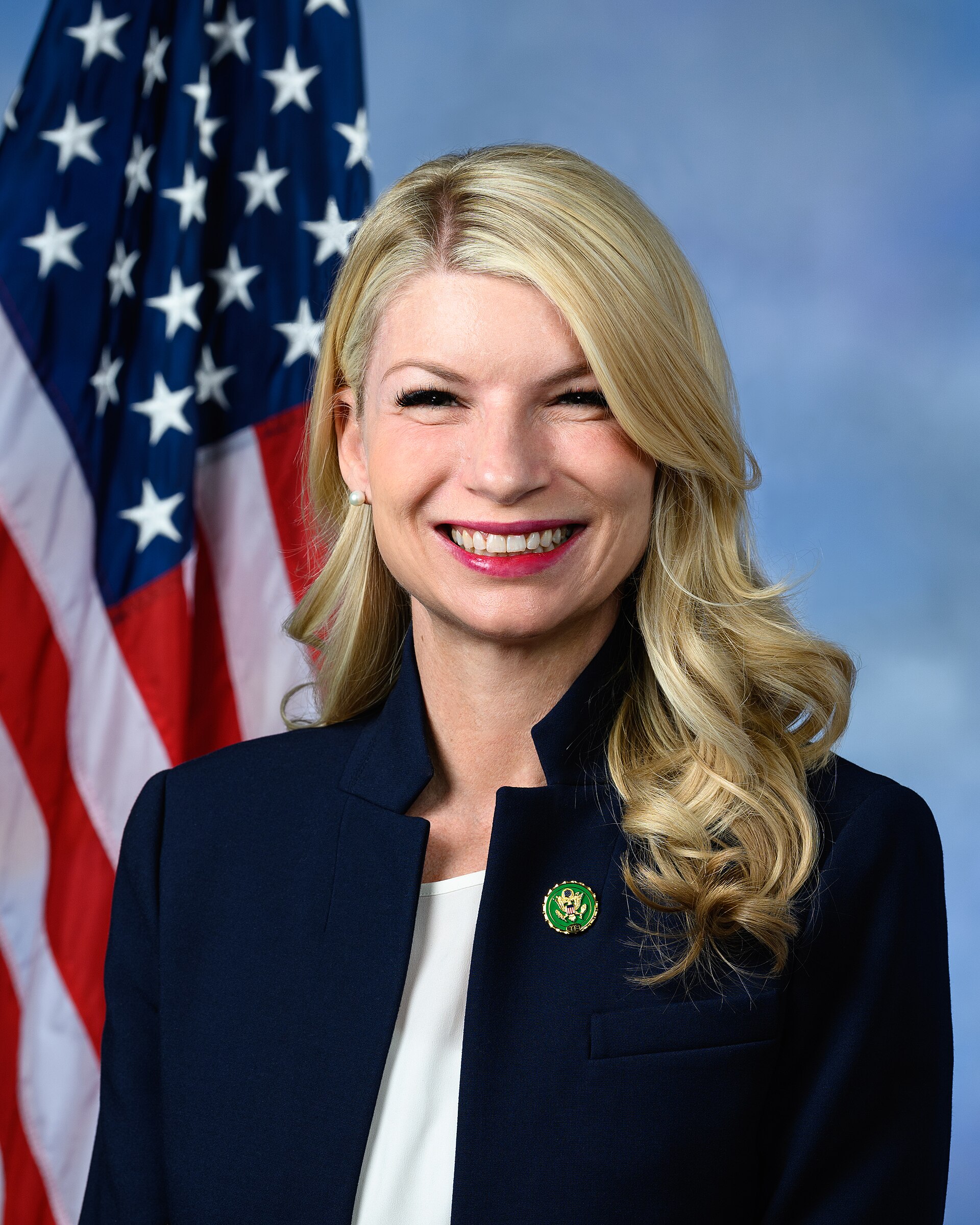 A smiling woman with long blonde hair, wearing a dark blue blazer and a white top, with a US flag behind her.