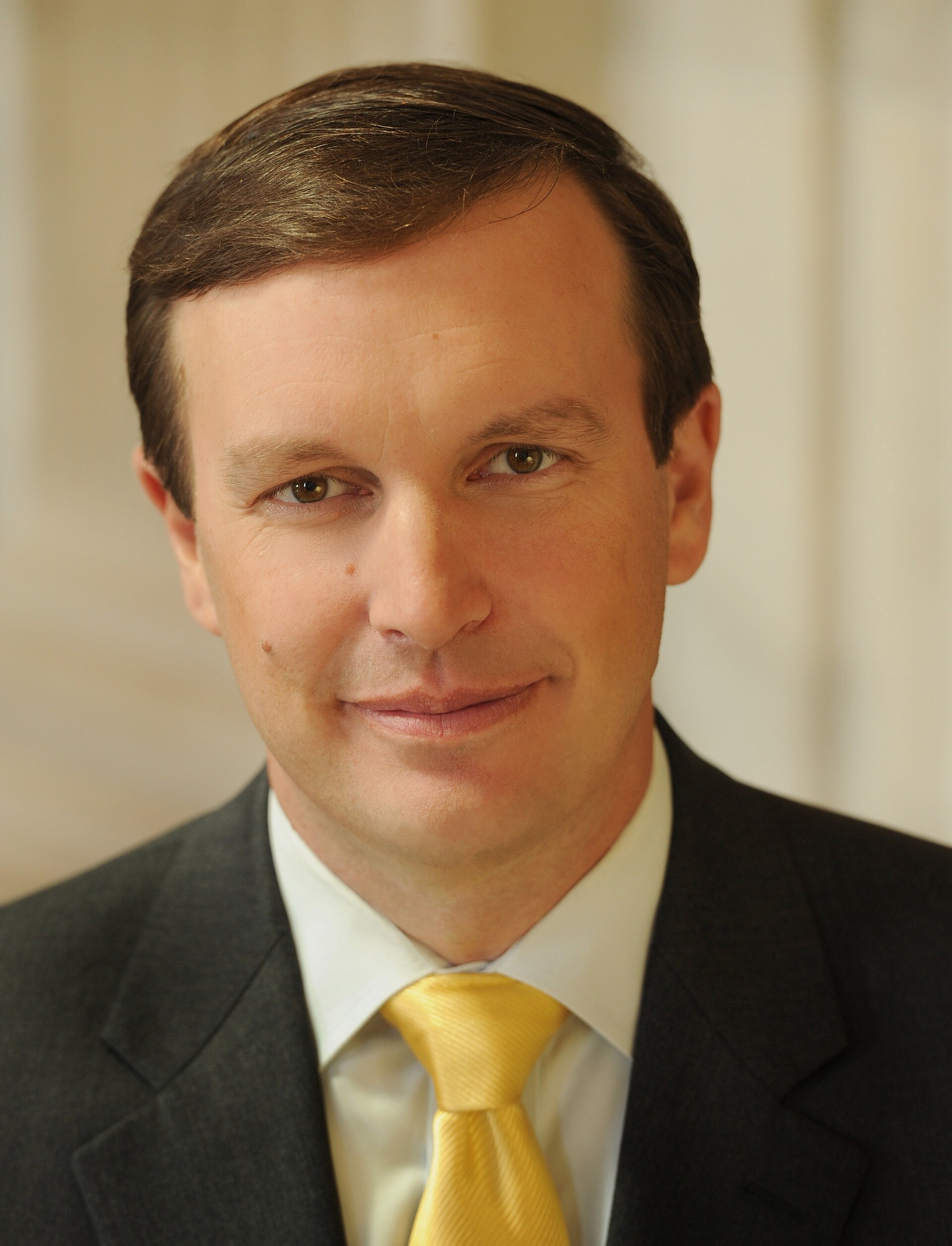 A man in a dark suit, white shirt, and yellow tie smiles slightly, showing his teeth.