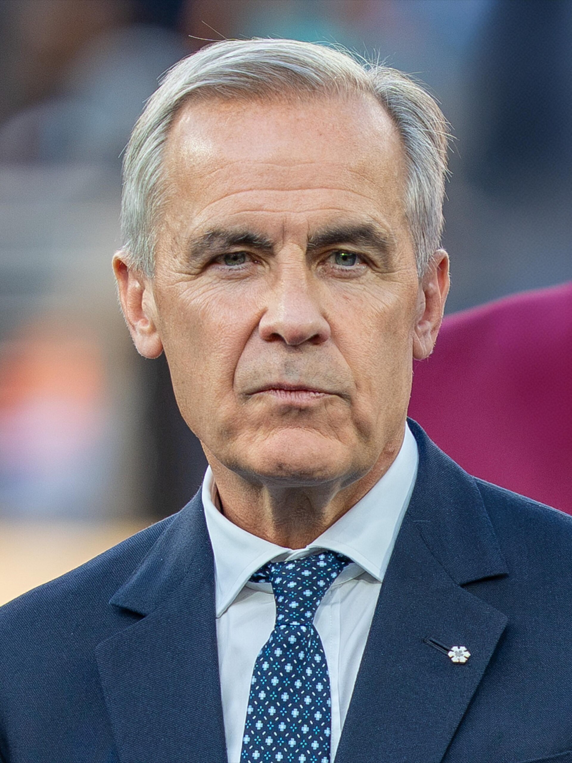 A middle-aged man with short gray hair, wearing a blue suit, white shirt, and blue tie with white dots, looking directly at the camera with a neutral expression.