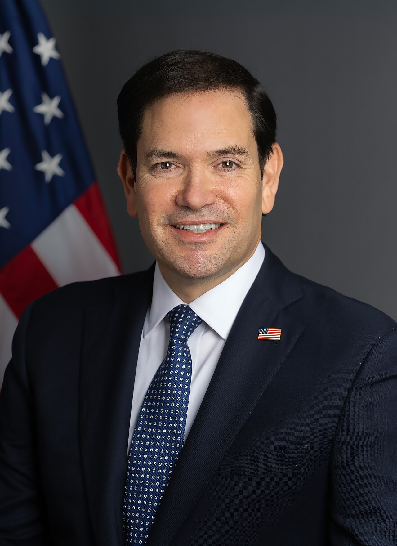 Marco Rubio in a suit and tie, with an American flag pin on his lapel, standing in front of a US flag.
