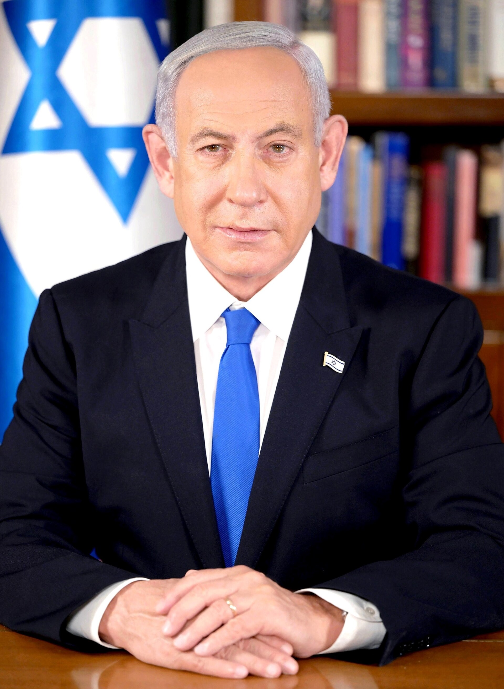 Benjamin Netanyahu, wearing a dark suit, white shirt, and blue tie, sits with his hands clasped on a wooden table. Behind him, to his left, is the Israeli flag, and to his right are bookshelves filled with books.