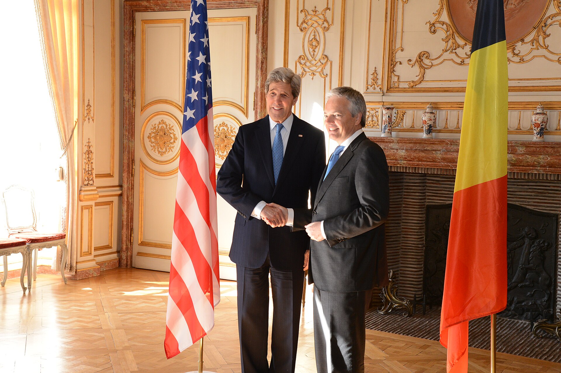 Two men in suits shake hands in front of the American flag and the Belgian flag.