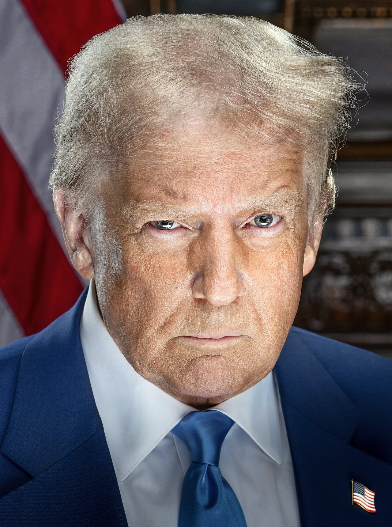 Donald Trump, a white man with blond hair, wearing a blue suit, white shirt, and blue tie, with an American flag pin on his lapel, looking directly at the camera with a serious expression.