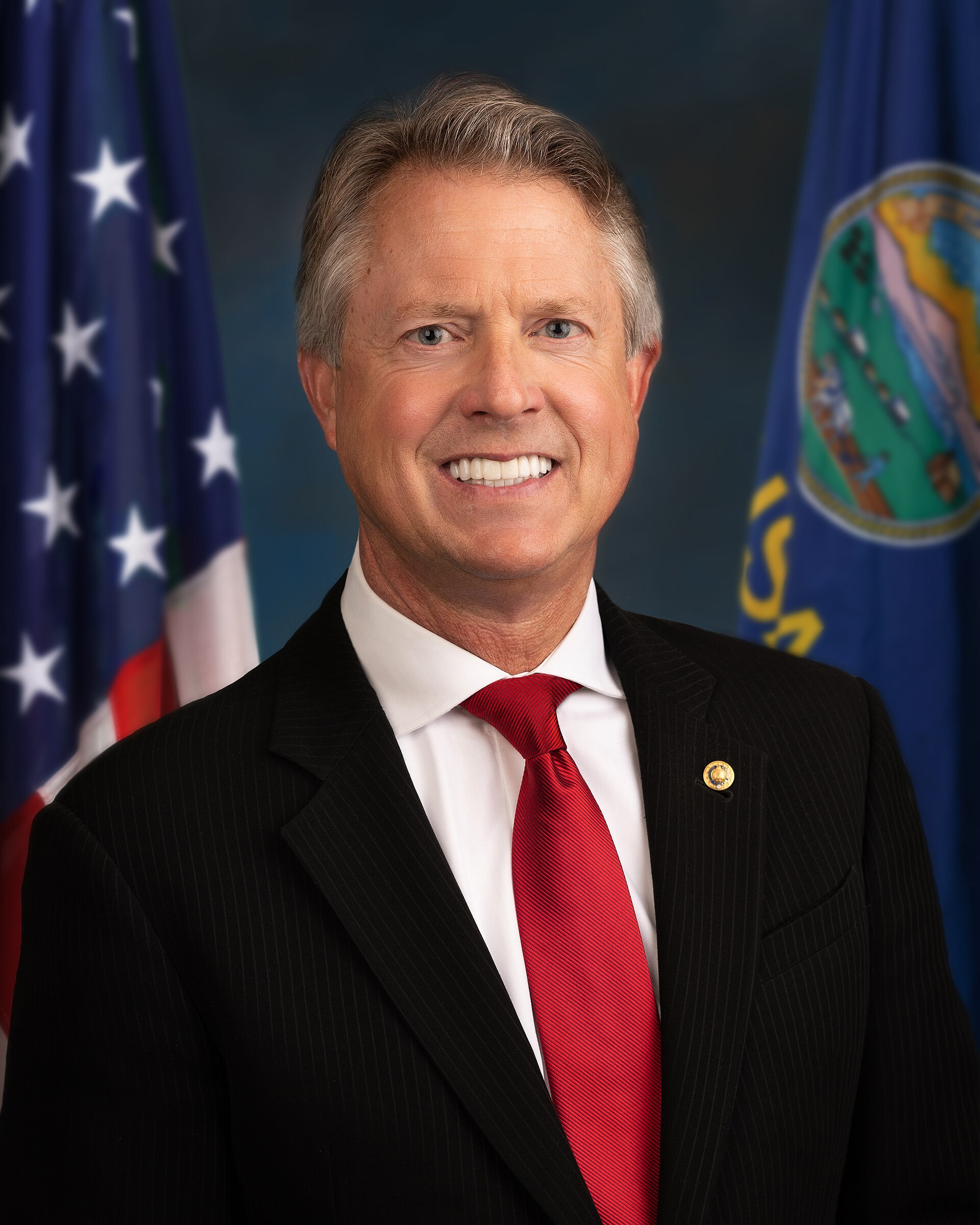 A middle-aged man with short gray hair, wearing a black suit, white shirt, and red tie, with a pin on his lapel, stands in front of two flags.