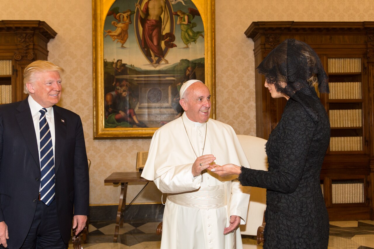 Donald Trump in a black suit and blue tie, smiling, standing next to Pope Francis in white robes and a woman in a black dress and veil, in a room with a large painting and bookshelves.