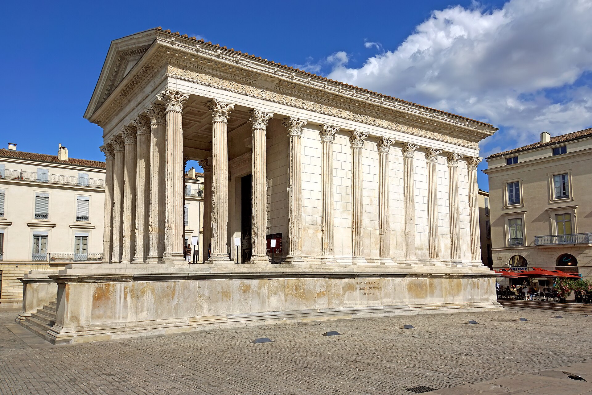 A wide shot of the Maison Carrée, a Roman temple in Nîmes, France, with its neoclassical facade and Corinthian columns. The temple is situated in a paved square with a blue sky and clouds overhead. Adjacent buildings and an outdoor cafe are visible to the right.