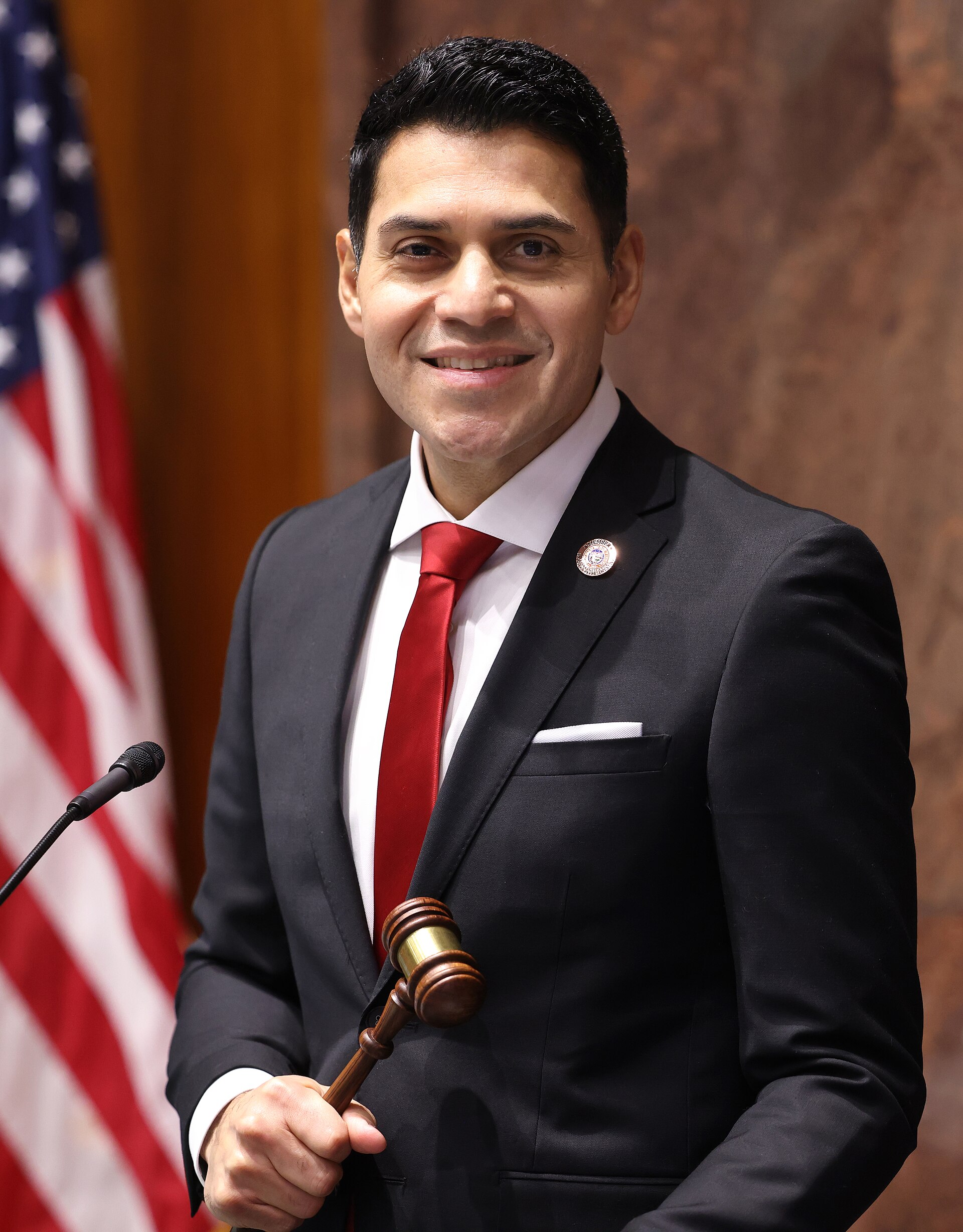 House Speaker Steve Montenegro, a man with short black hair, wearing a black suit, white shirt, and red tie, holding a gavel and standing in front of an American flag.