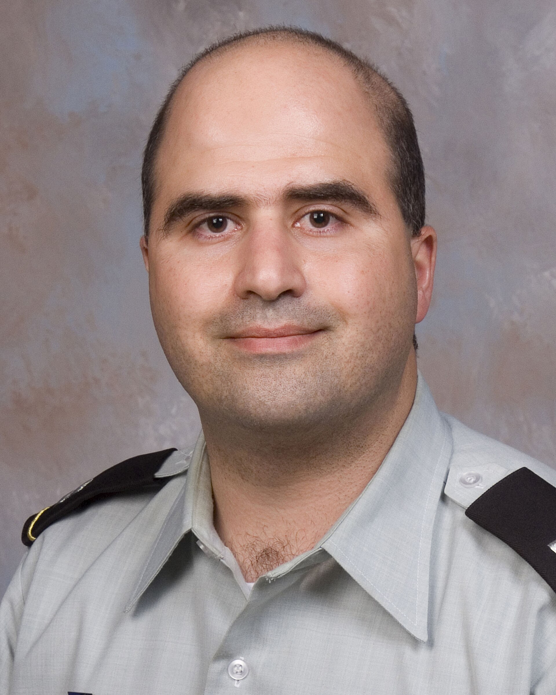 A head and shoulders portrait of a middle-aged man in a military uniform with a gray shirt and black epaulets, against a mottled gray background.