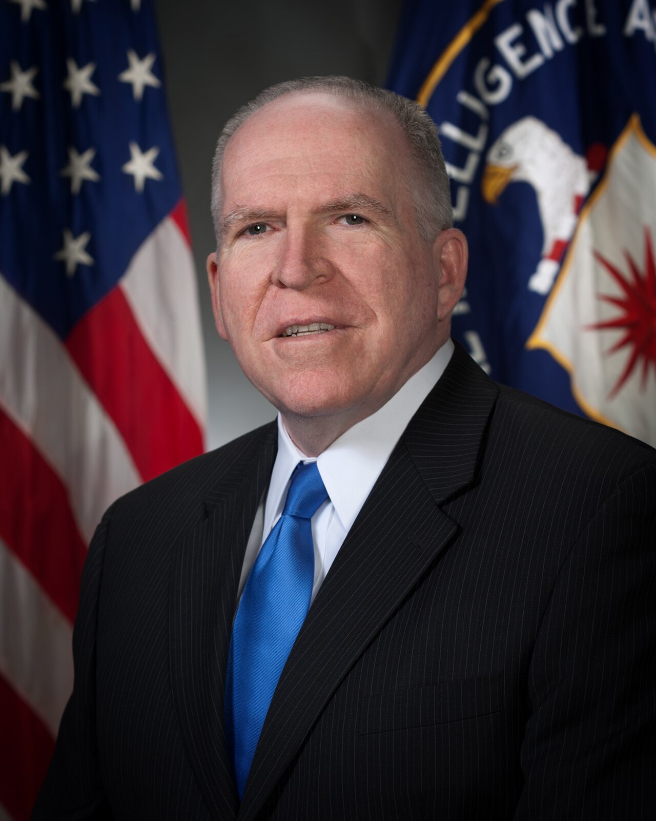 John Brennan, former CIA Director, in a suit and blue tie, stands in front of the American flag and the CIA seal.