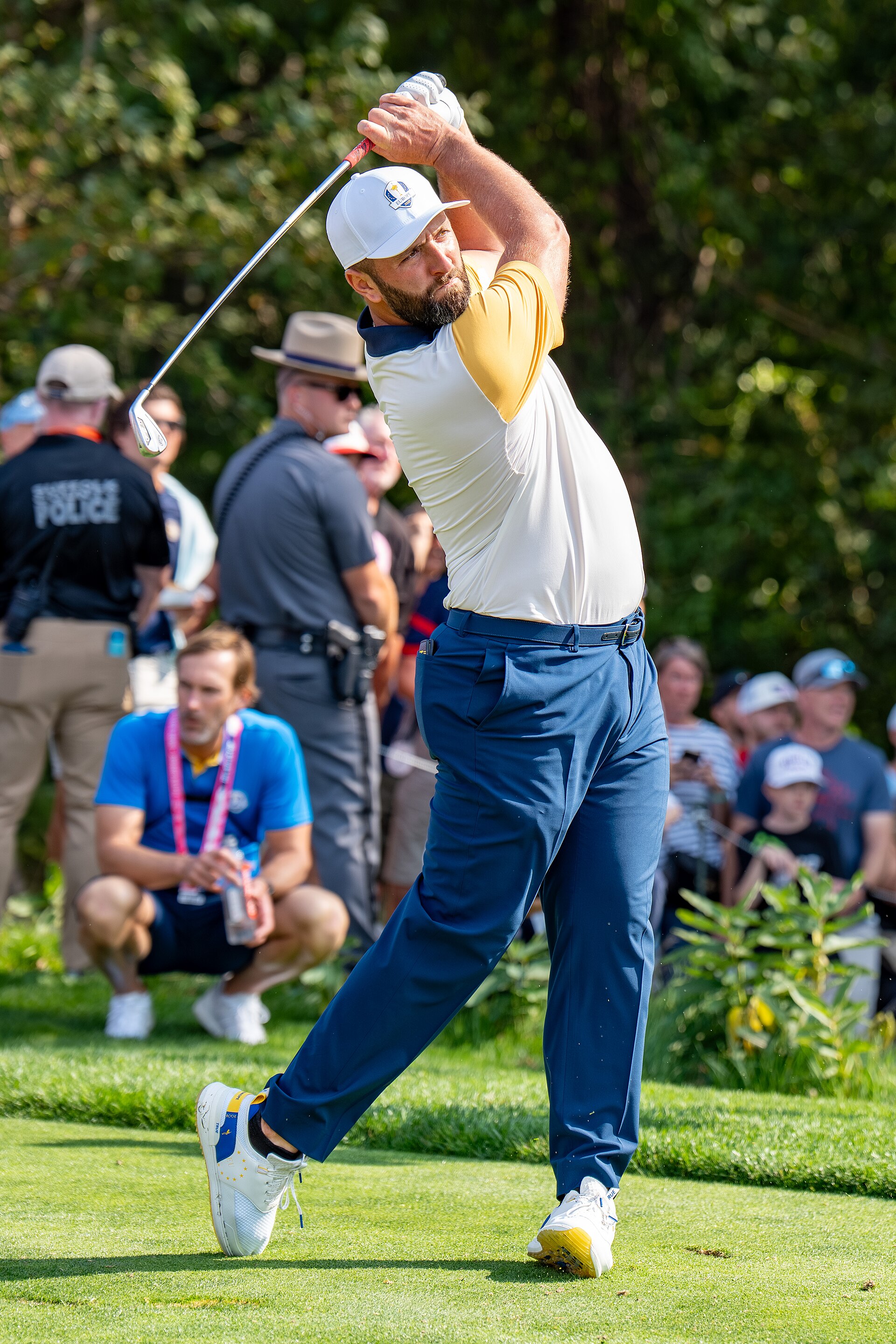 Jon Rahm swings a golf club during a tournament, wearing a white cap with a European emblem, a white and yellow polo shirt, and blue pants.