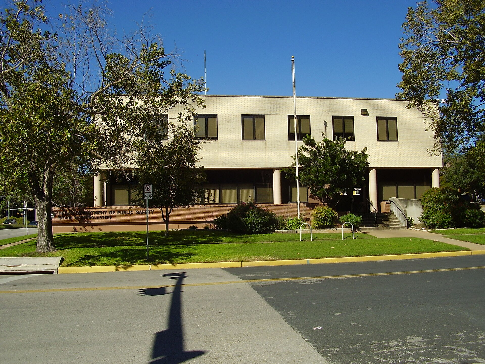 A light-colored brick building with dark windows, identified by signage as the Texas Department of Public Safety Headquarters, stands under a clear blue sky. Trees with green leaves partially obscure the building, and a shadow of a street sign is cast on the road in the foreground.