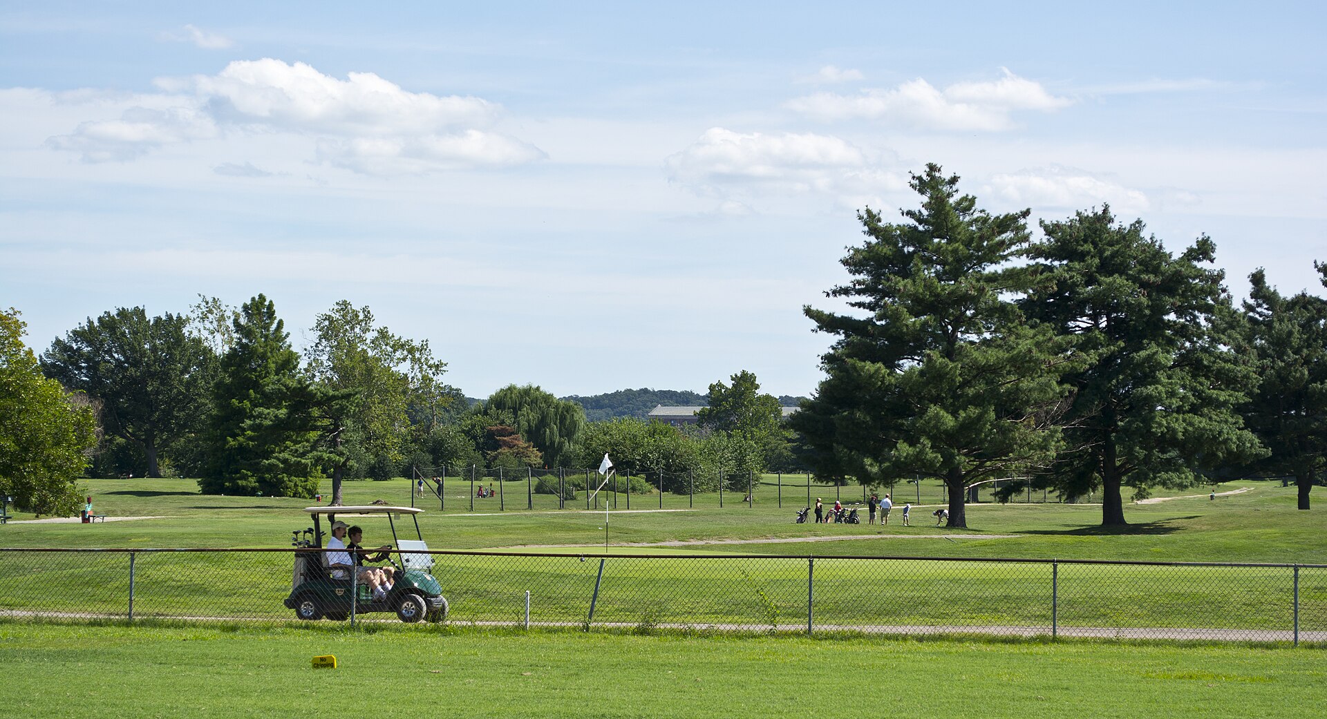 A golf course with several people playing golf and a golf cart driving on the grass.