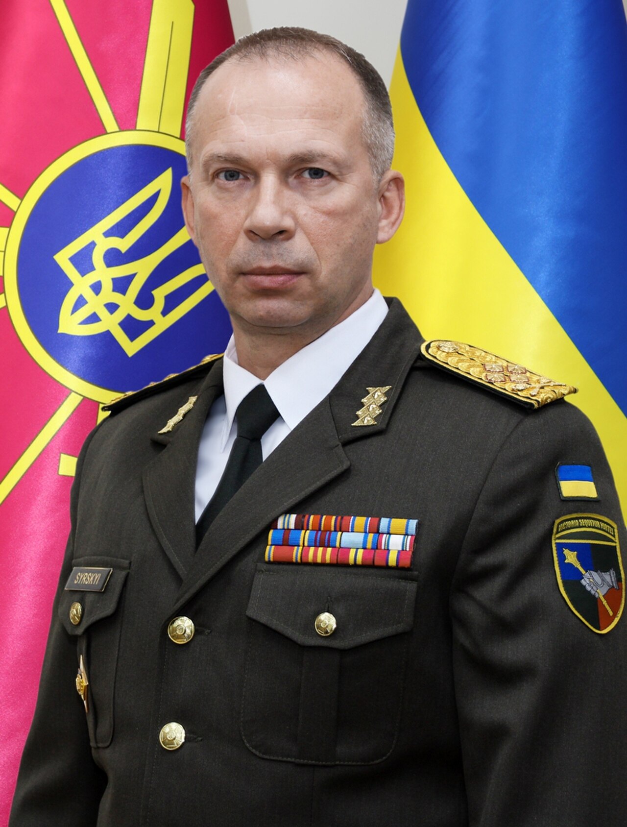 A man in a military uniform with medals and insignia stands in front of two flags.
