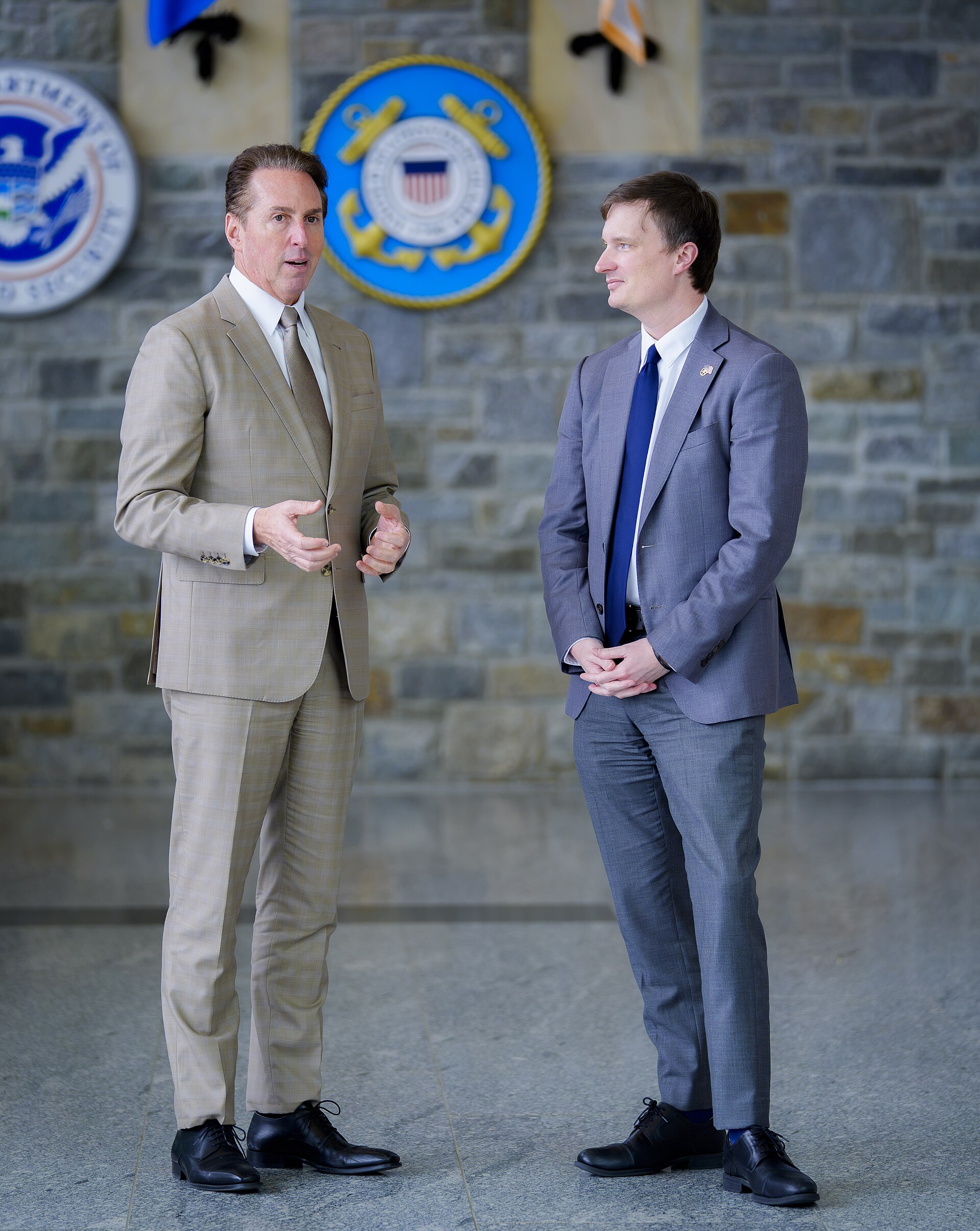 Two men in suits stand in front of stone walls with circular emblems.