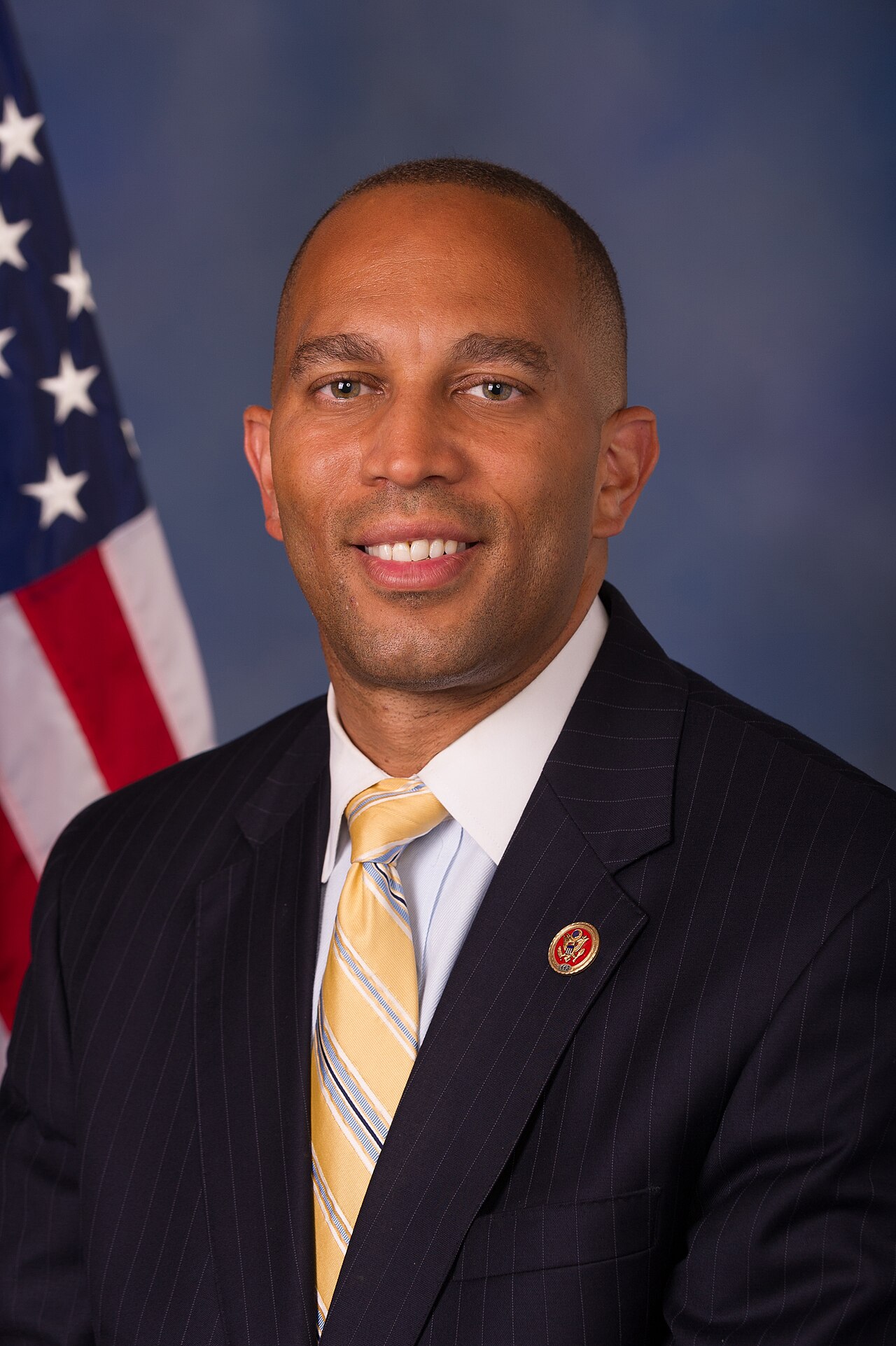 Hakeem Jeffries, wearing a dark suit, light blue shirt, and yellow tie, with an American flag behind him.