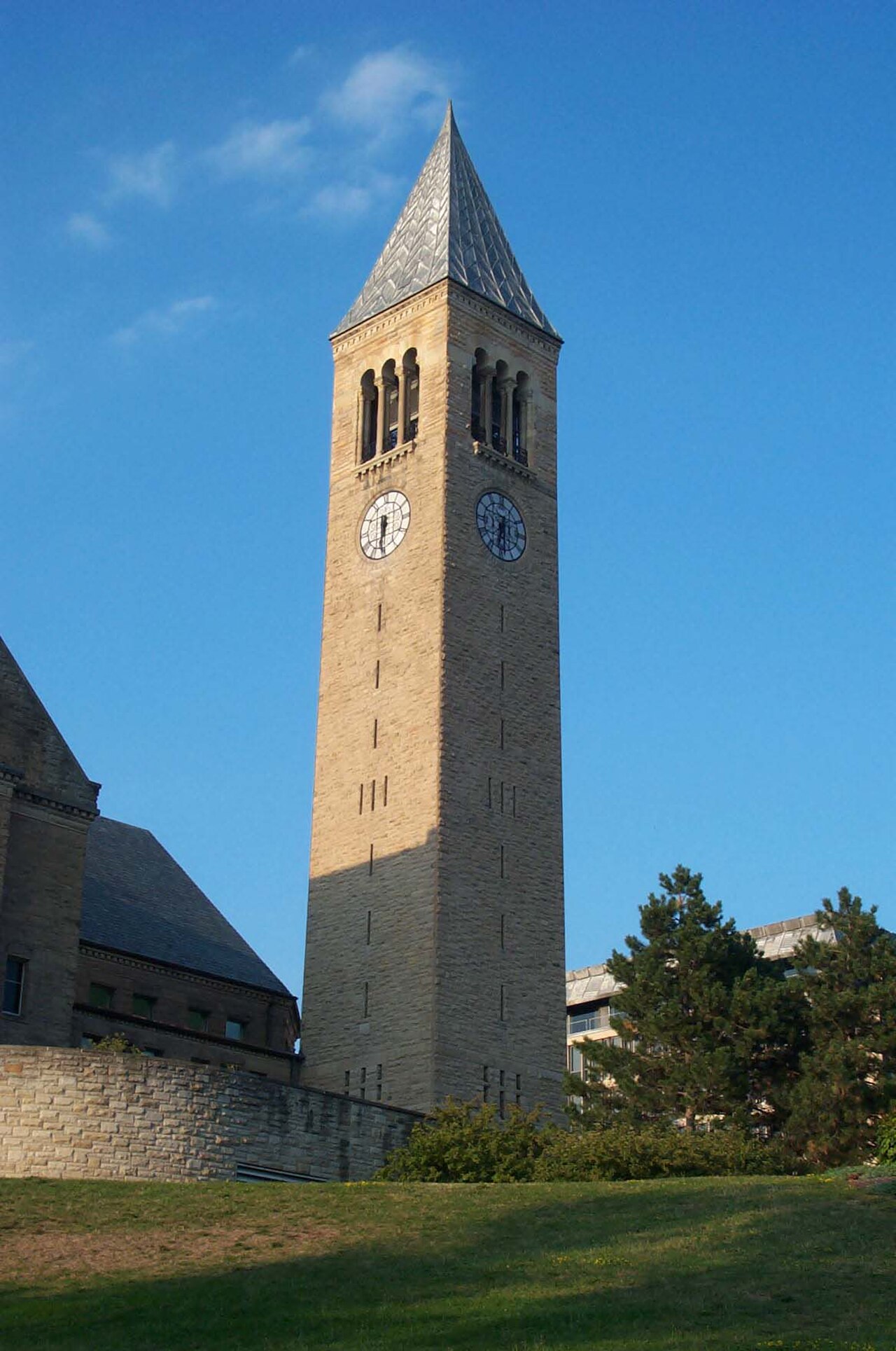 A tall, tan brick clock tower with a gray roof and two white clock faces, surrounded by green grass and trees, with a blue sky in the background.