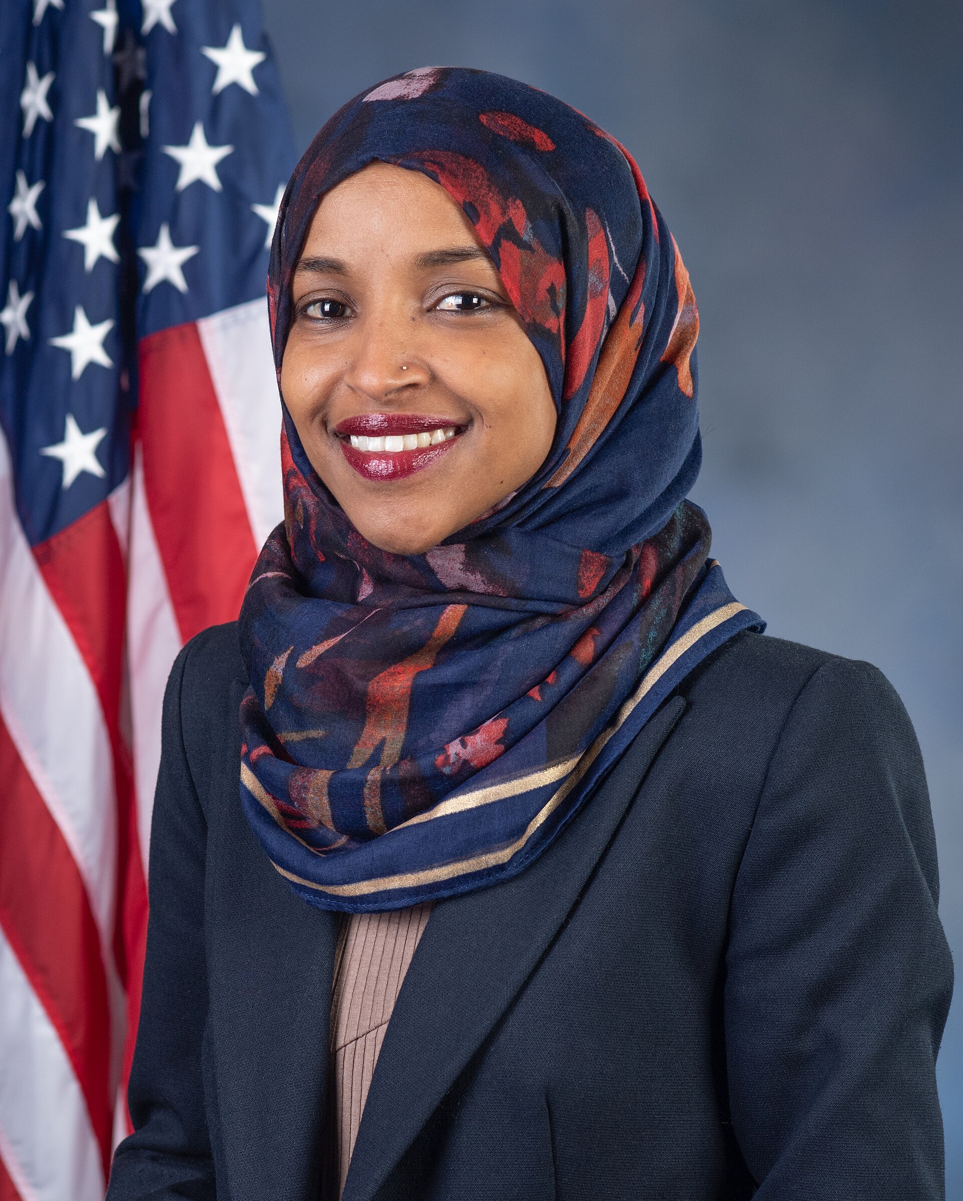 Ilhan Omar smiling and wearing a blue hijab with red accents, a dark blazer, and a tan top, standing in front of a US flag.