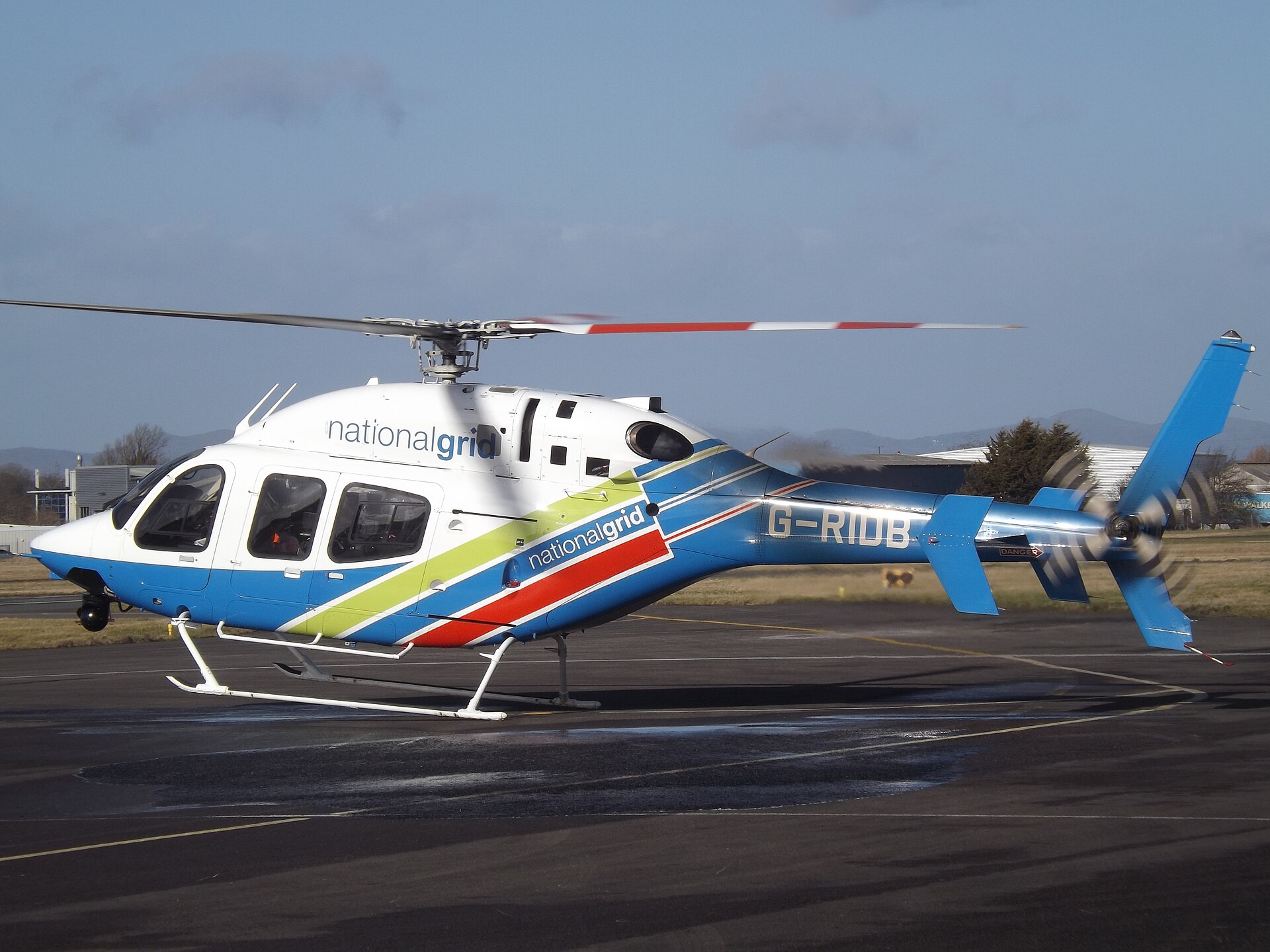 A white helicopter with blue, green, and red stripes and the text "nationalgrid" and "G-RIDB" is on an asphalt surface.