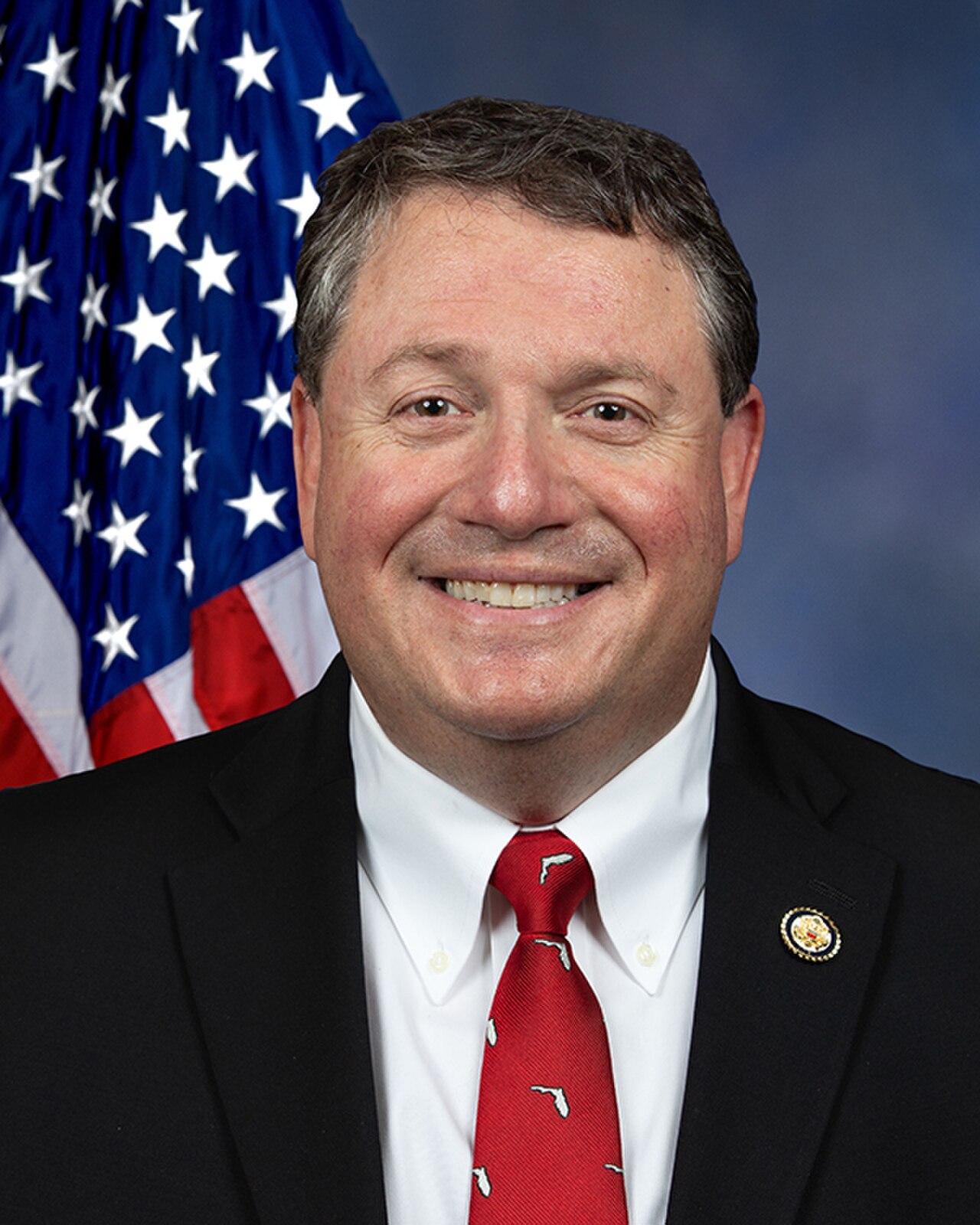 A smiling middle-aged man in a black suit, white shirt, and red tie with a Florida pattern, standing in front of an American flag.