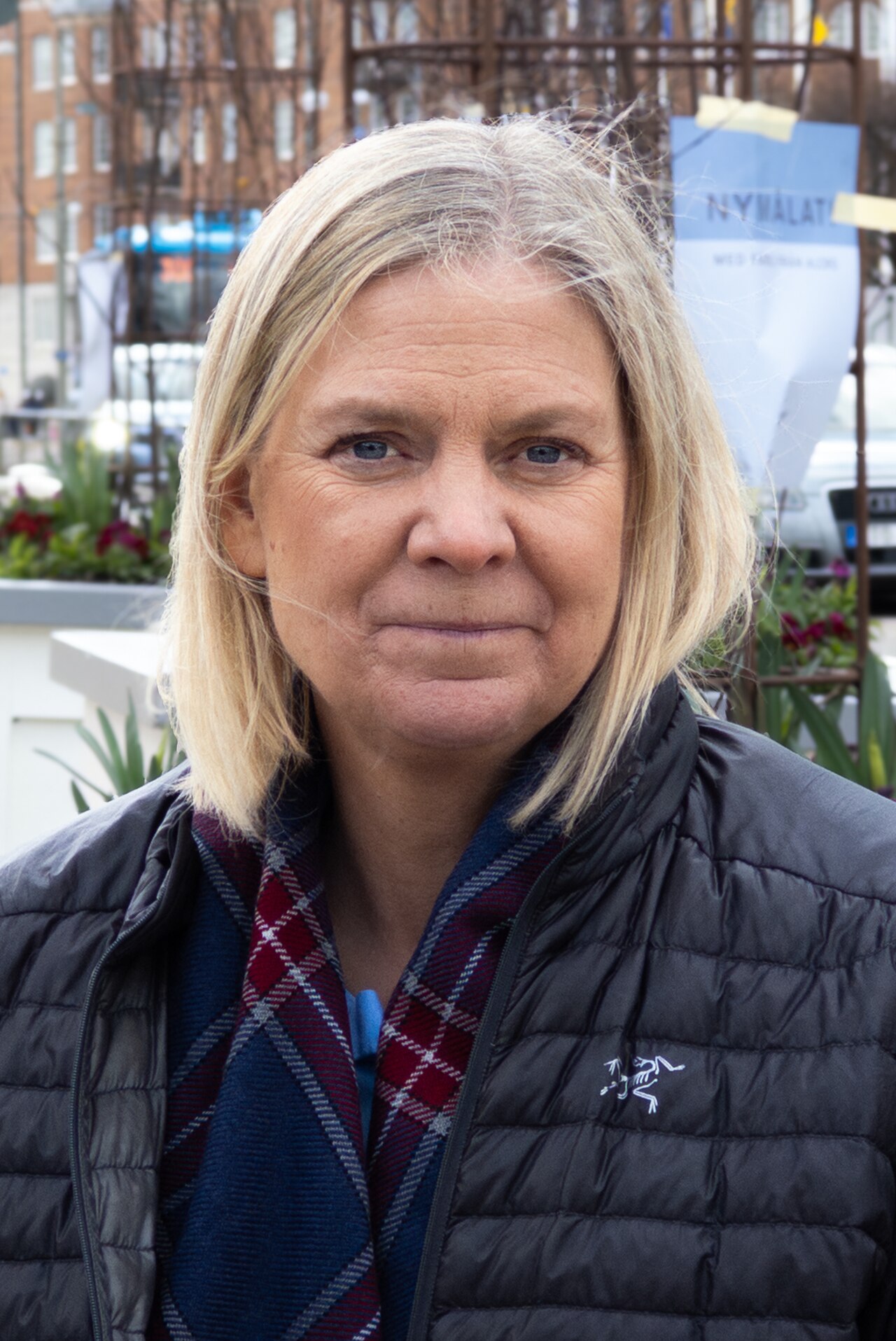 A woman with shoulder-length blonde hair, blue eyes, and a neutral expression, wearing a black puffer jacket and a red and blue plaid scarf.
