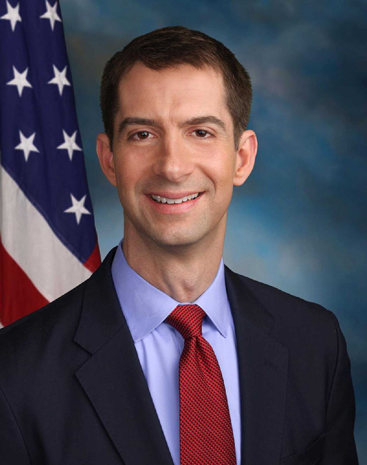 Tom Cotton, a middle-aged white man with short brown hair, wearing a dark suit, light blue shirt, and red tie, stands in front of an American flag and blue background.