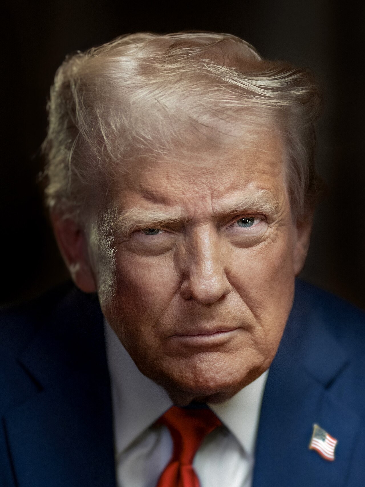 Donald Trump in a dark suit, white shirt and red tie with a flag pin, looking into the camera against a dark background.