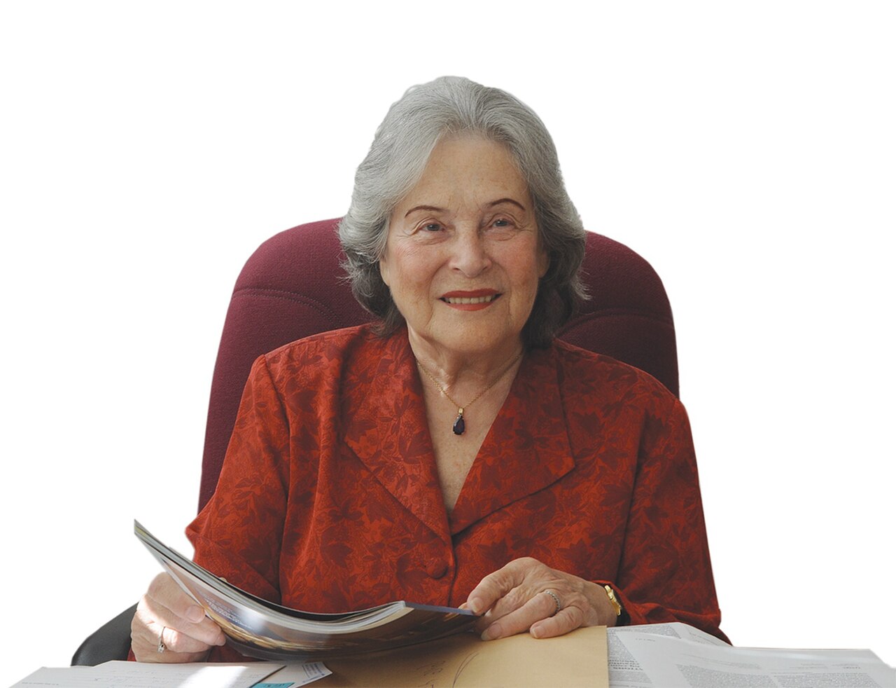 A middle-aged woman with short gray hair, wearing a red blouse and a necklace, sitting in a red chair at a desk with papers, looking at the camera.