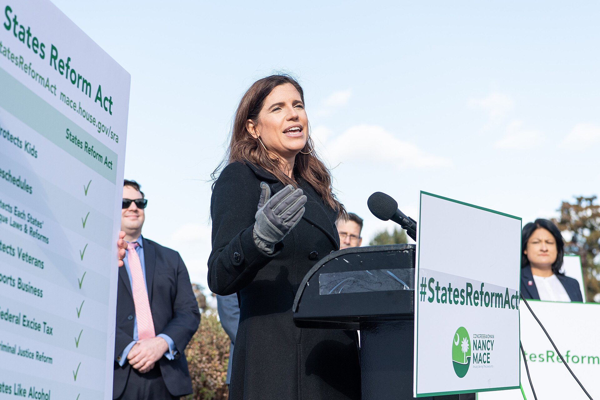 A woman in a black coat and grey gloves speaks at a podium with a microphone. A sign behind her reads "#StatesReformAct" and "CONGRESSWOMAN NANCY MACE".