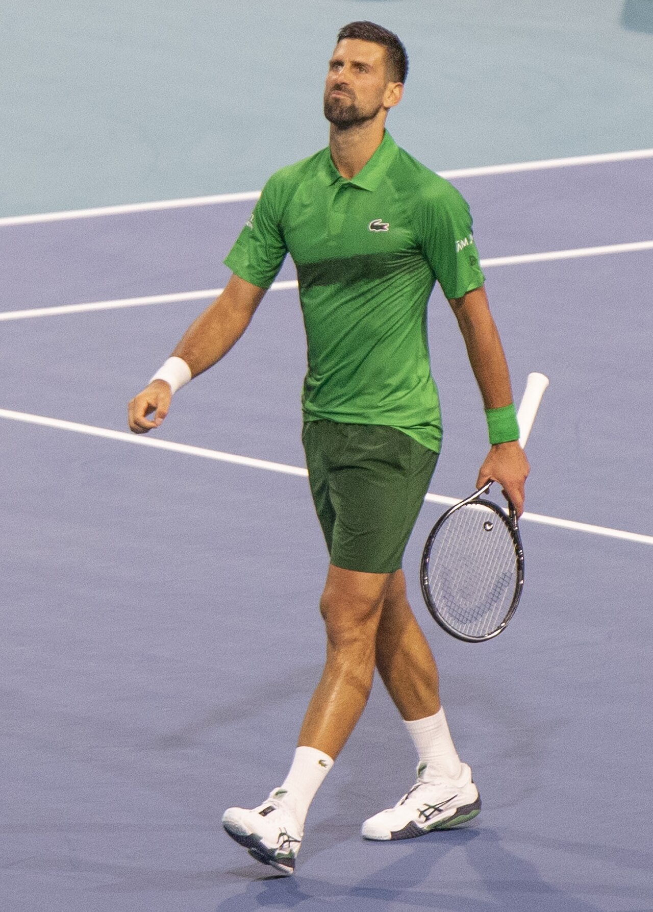 A man with short dark hair wearing a green shirt and shorts holding a tennis racket on a tennis court