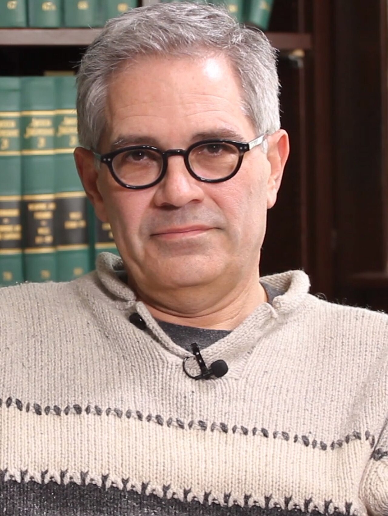 A man with short gray hair, wearing black-framed glasses and a beige sweater with dark stripes, appears in front of a bookshelf.