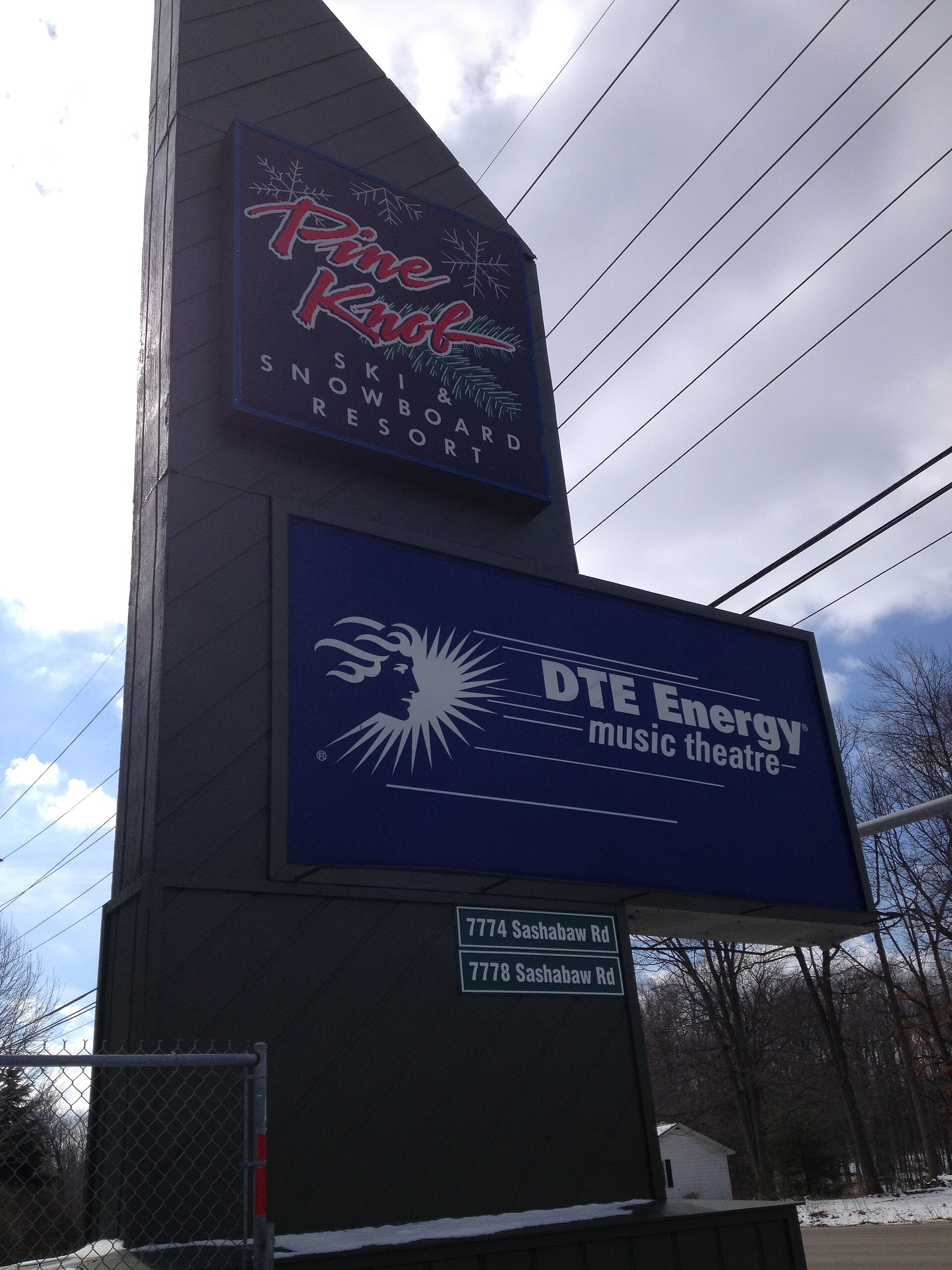 A tall signpost with two displays. The top display reads 'Pine Knob Ski & Snowboard Resort'. The bottom display reads 'DTE Energy music theatre' with a logo of a sunburst and a profile of a face. Below the displays are two green signs with white text indicating addresses: '7774 Sashabaw Rd' and '777