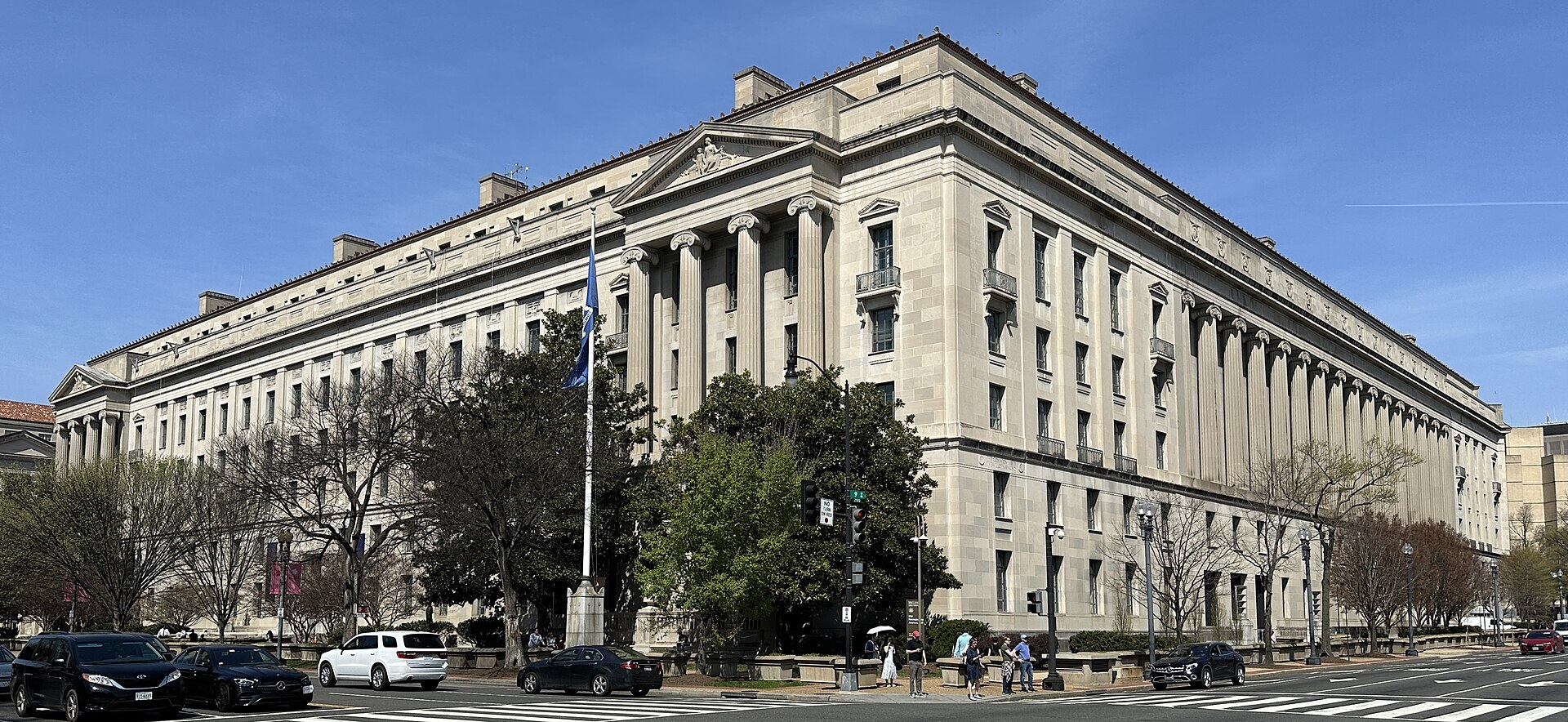 The exterior of the Department of Justice building in Washington D.C. on a sunny day, with cars and pedestrians visible on the street in front.