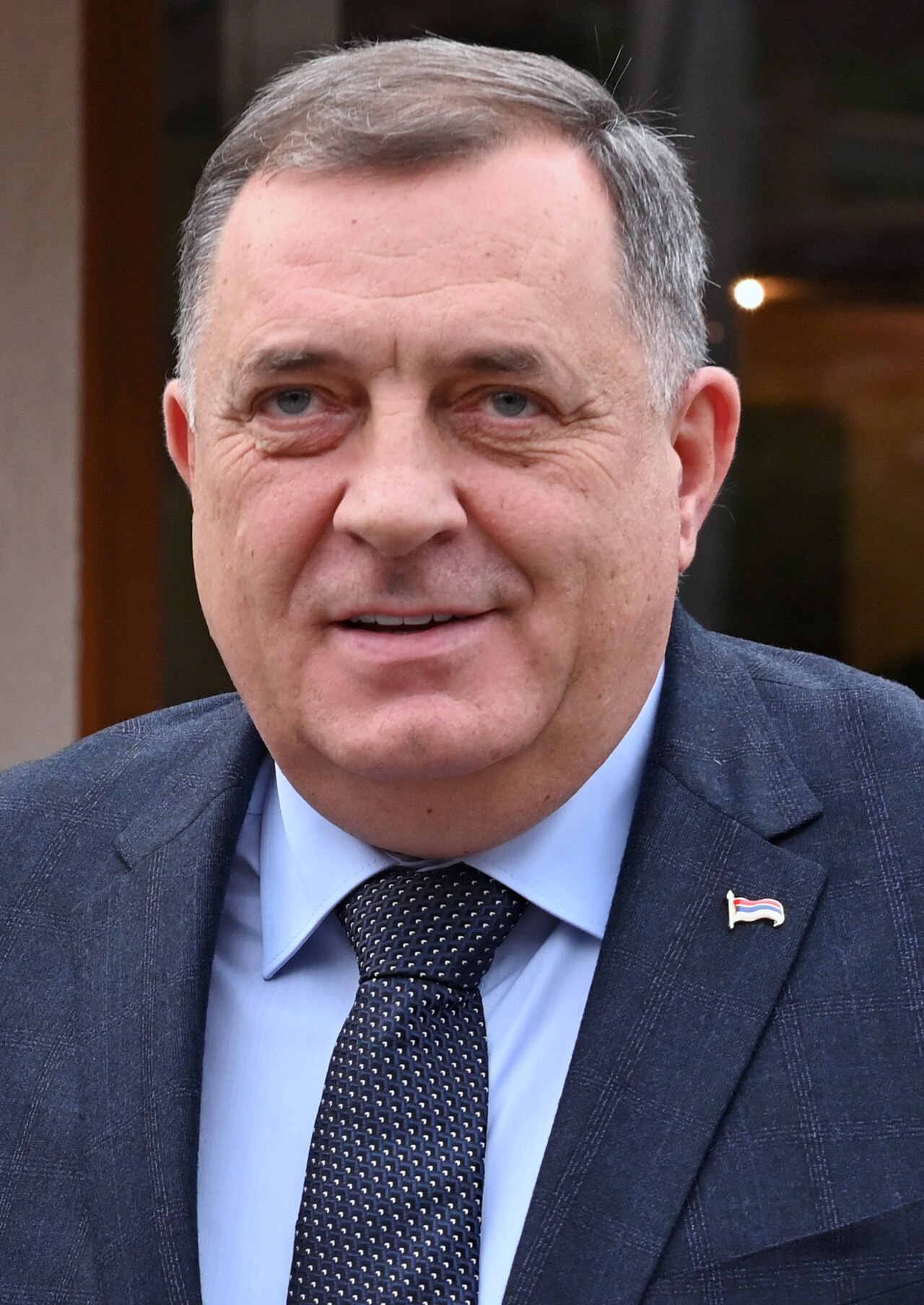 Milorad Dodik, wearing a dark blue suit, light blue shirt, and dark blue tie with white dots, and a small flag pin on his left lapel.