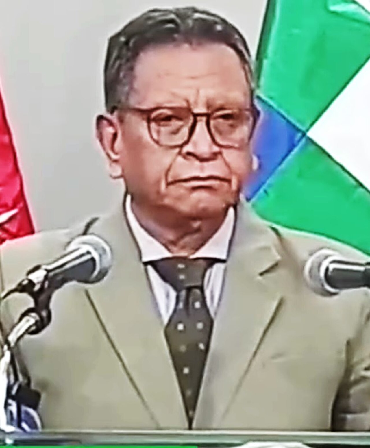 A man with short dark hair, wearing a tan suit, white shirt, dark tie with light dots, and glasses, stands at a podium with two microphones, in front of a Bolivian flag.