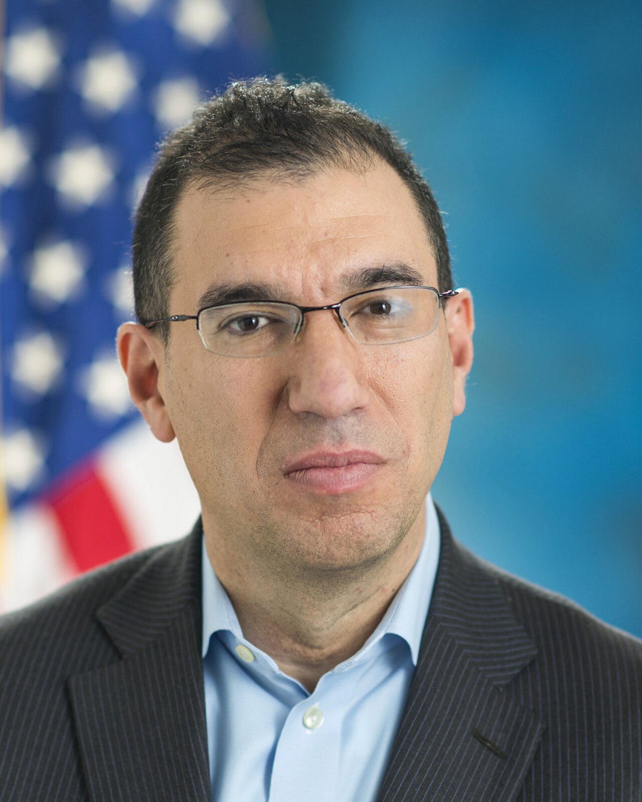 A man with short dark hair, wearing glasses, a dark suit jacket, and a light blue collared shirt, standing in front of a blurred American flag and blue background.