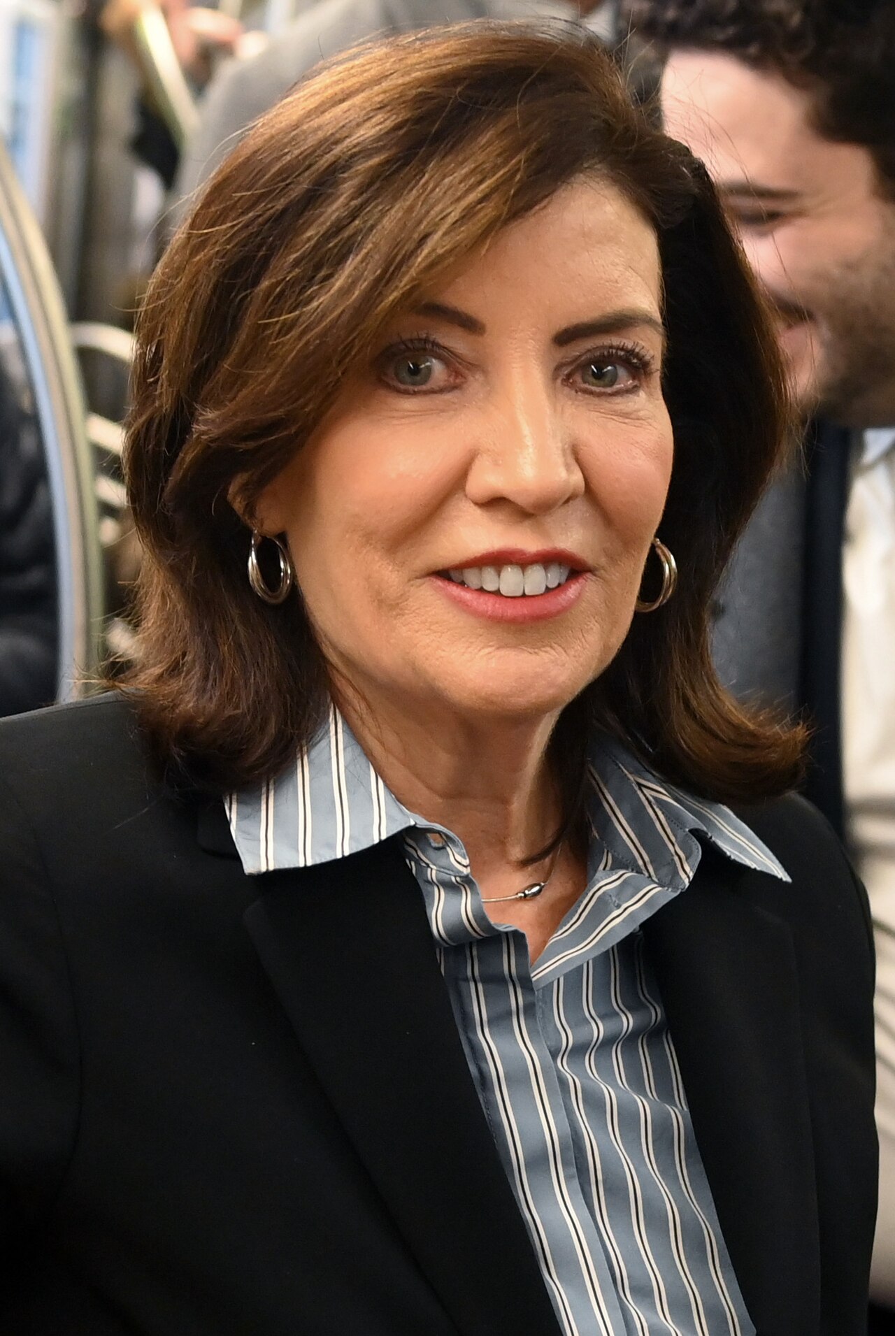 A smiling middle-aged woman with brown hair wearing a black blazer and a blue and white striped collared shirt.