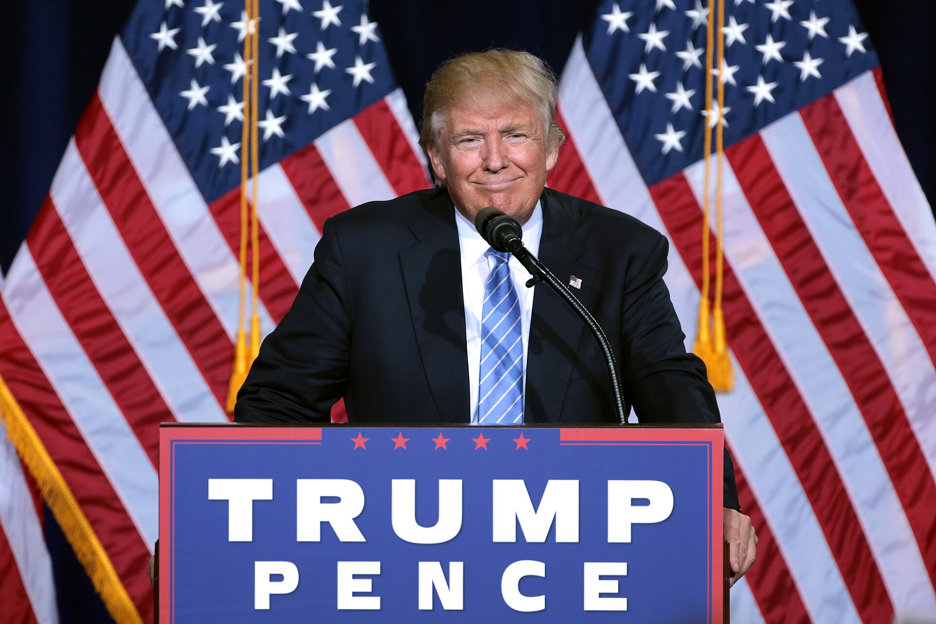 Donald Trump at a podium with a sign that reads TRUMP PENCE