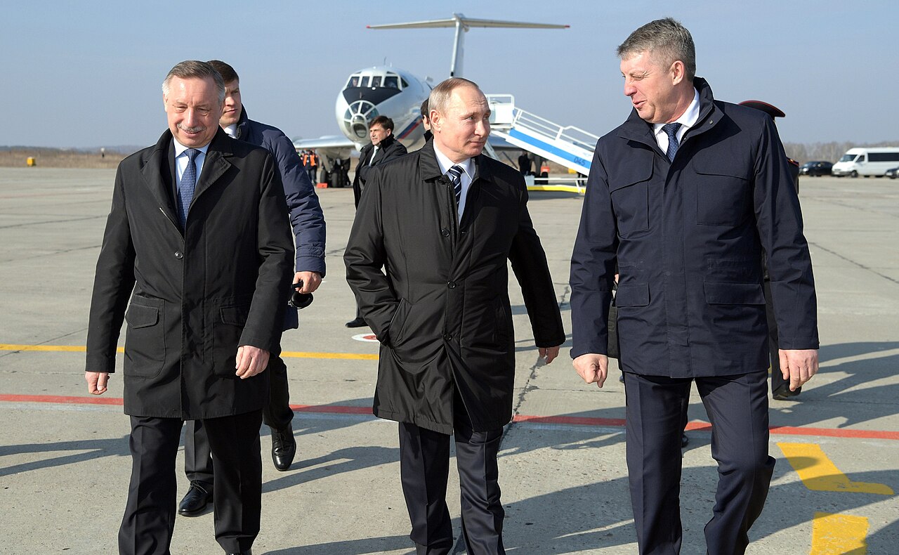 Three men in dark coats walk on an airport tarmac with a plane in the background.