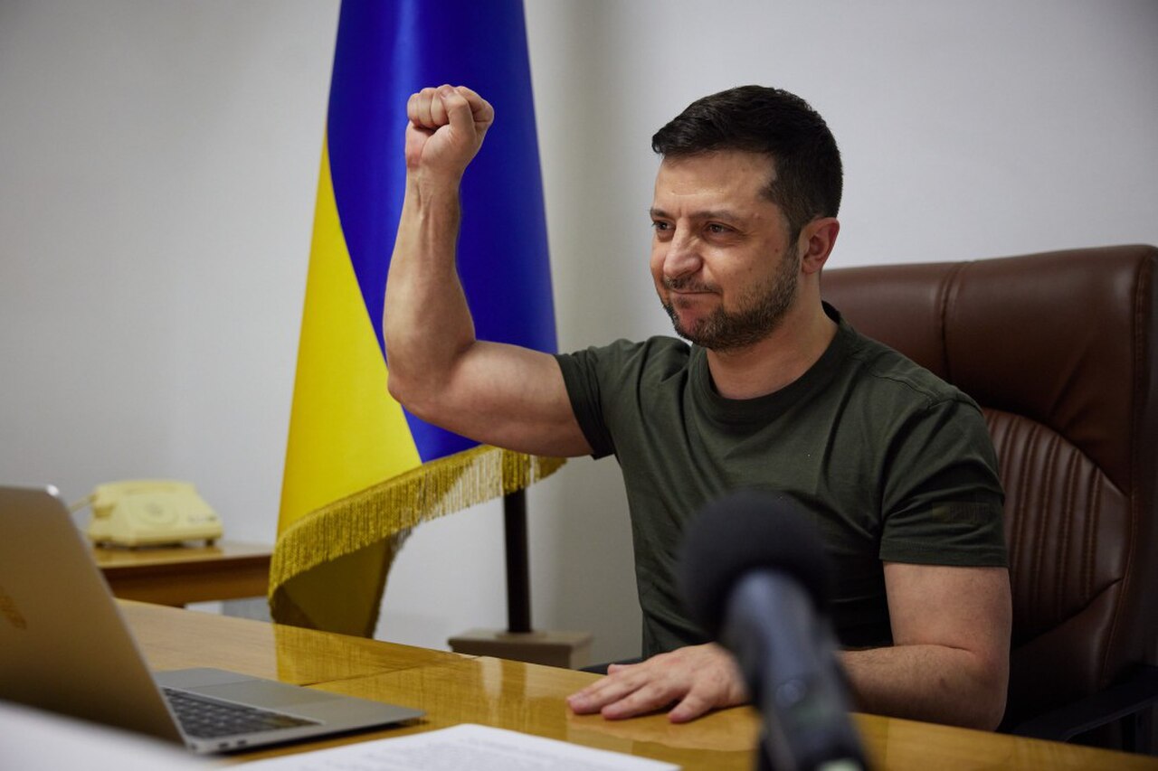 A man with short dark hair and a beard, wearing a green t-shirt, sitting at a desk with a laptop and microphone in front of him, and a Ukrainian flag behind him, with his right fist raised.