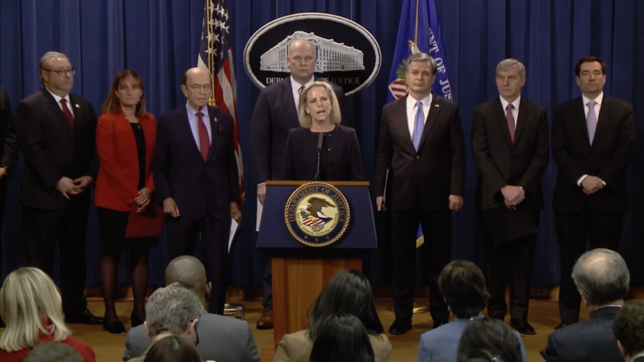 A group of people stand behind a podium with the Department of Justice seal. A woman is speaking at the podium. Several men and one woman stand behind her. The US flag and another flag are visible.
