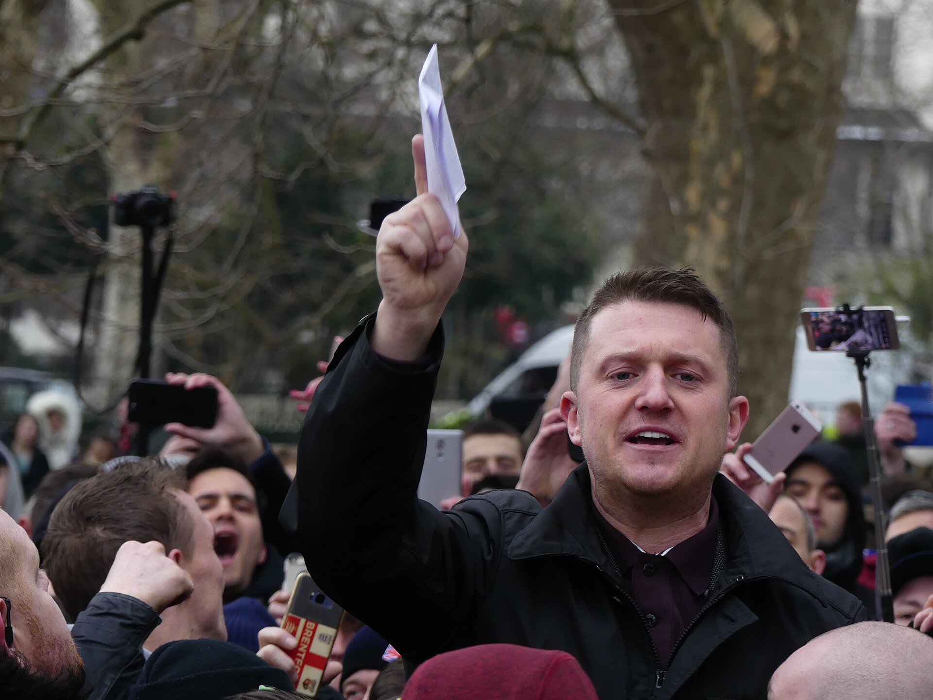 A man in a black jacket and maroon shirt, with short brown hair, raises his right hand holding a white piece of paper. He is surrounded by a crowd of people, many of whom are holding up their phones to take pictures or record videos. The man is looking towards the camera with his mouth slightly open