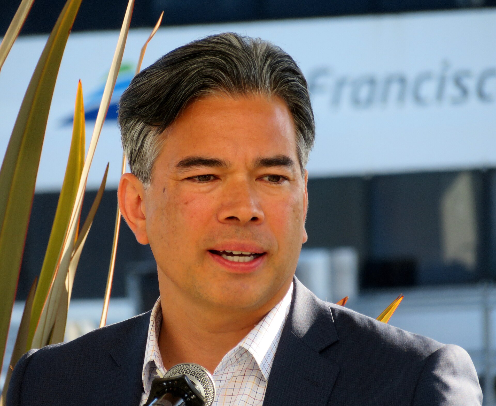 A man with short graying hair, wearing a dark suit jacket and white collared shirt, speaking into a microphone.