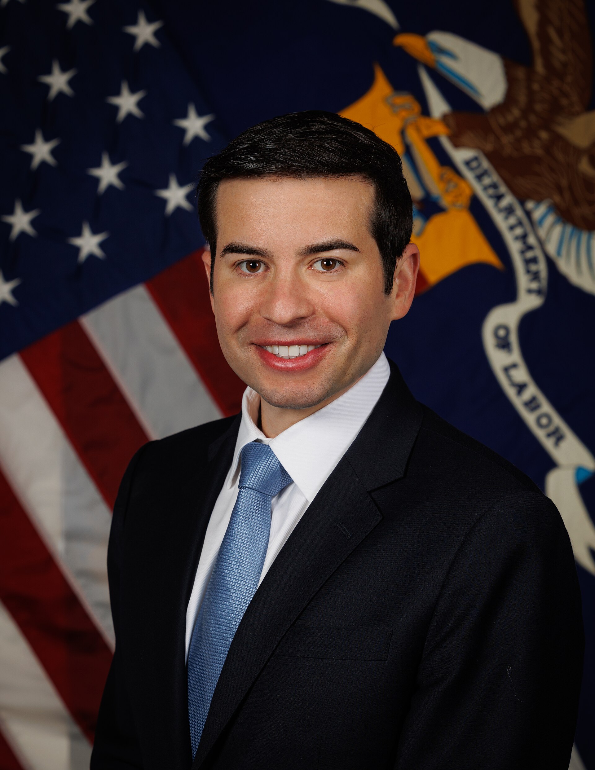 A smiling man in a suit and tie stands in front of an American flag and a Department of Labor flag.