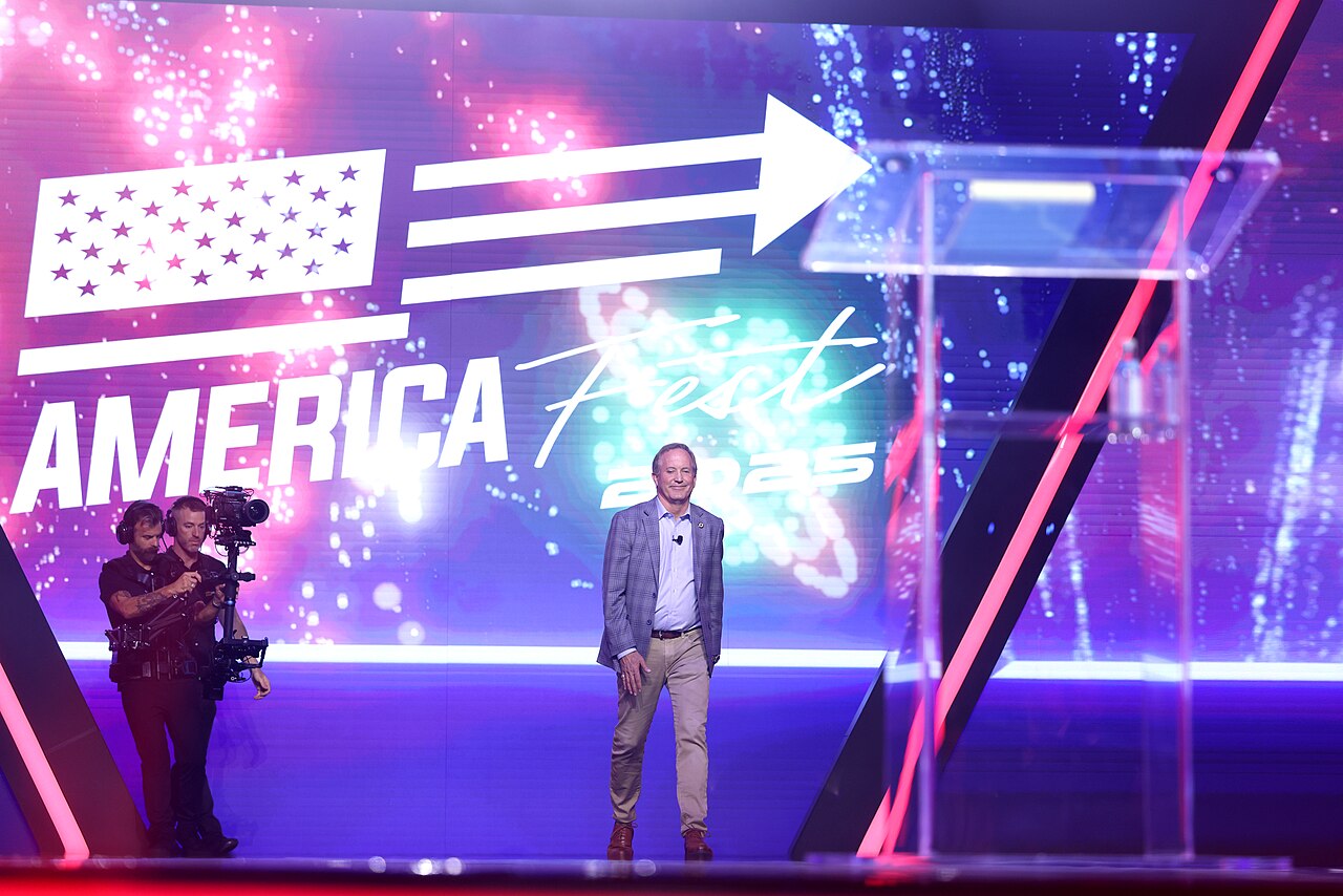 A man in a blue suit stands on a stage with a large screen behind him displaying a graphic with an American flag and the words 'AMERICA First' in white letters.