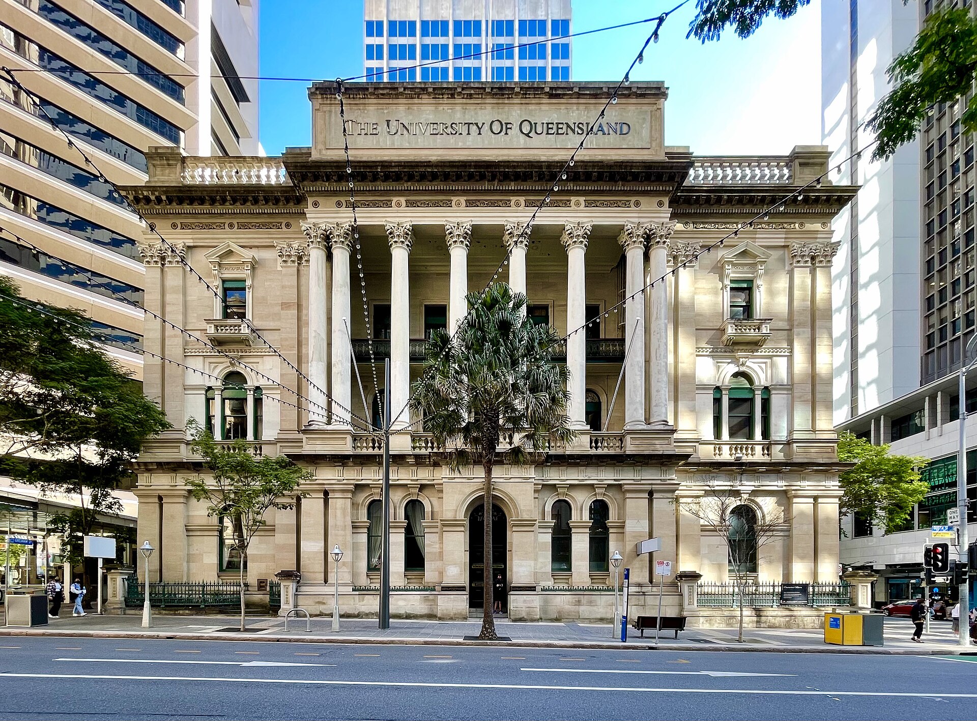 A grand, ornate building with columns and the words "THE UNIVERSITY OF QUEENSLAND" inscribed above the entrance, set against a backdrop of modern skyscrapers.