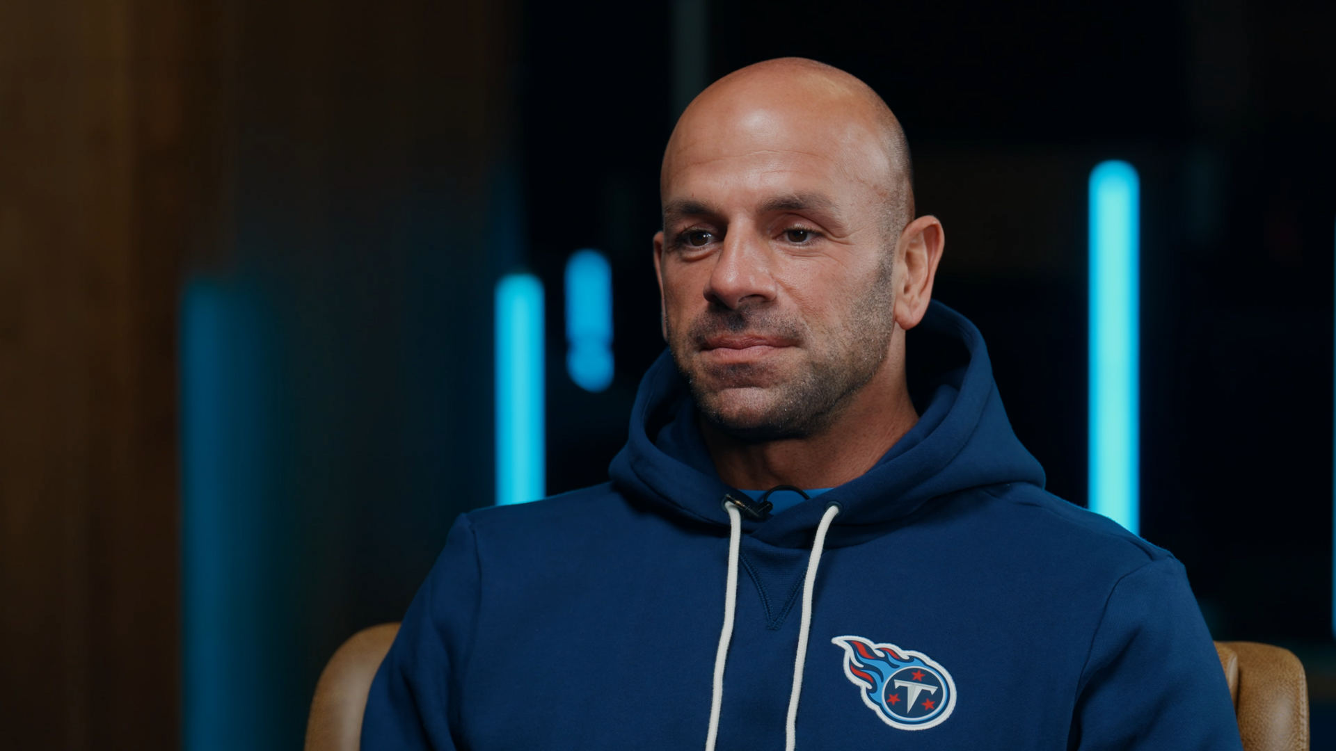 A bald man wearing a blue hoodie with the Tennessee Titans logo on it, looking down and to the side.